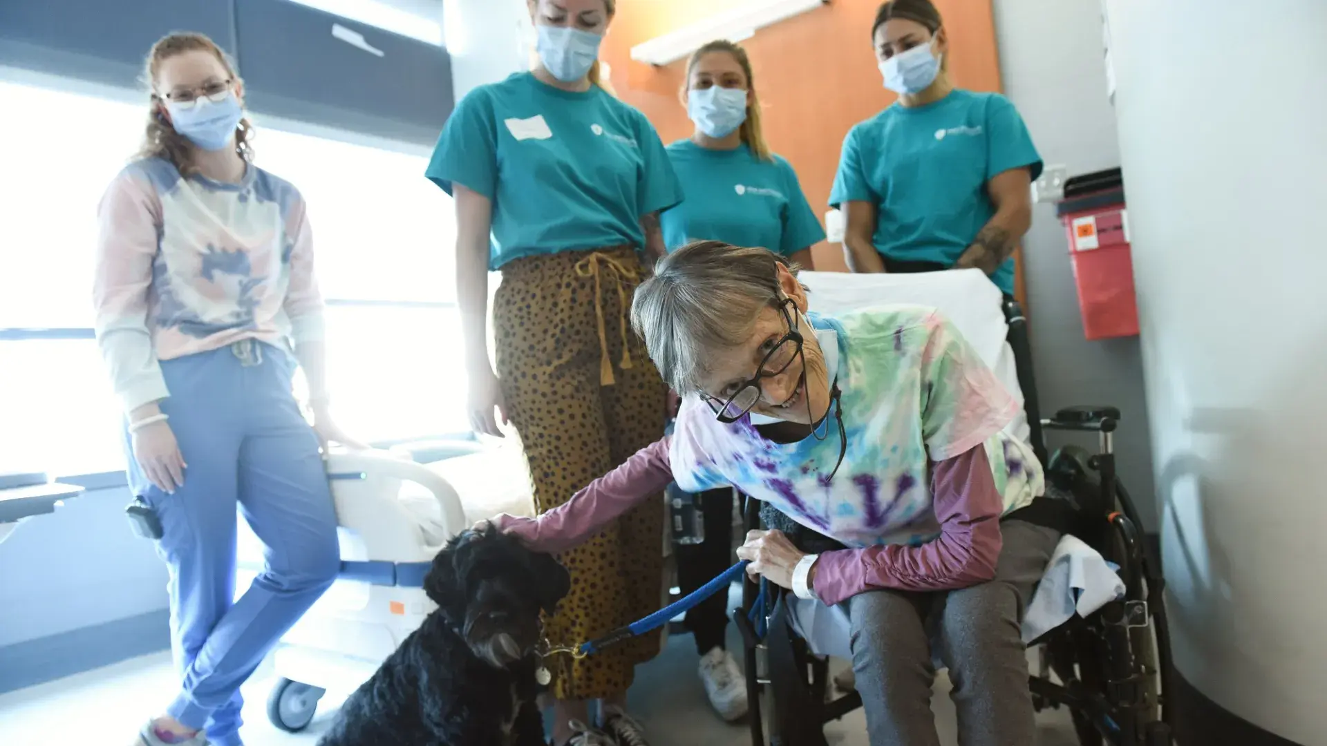 woman in wheelchair pets a dog while people in masks stand behind her