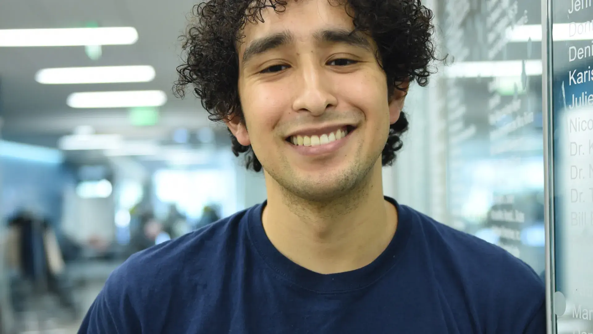 man with curly black hair and mgh ihp physical therapy on his tshirt leans against a wall