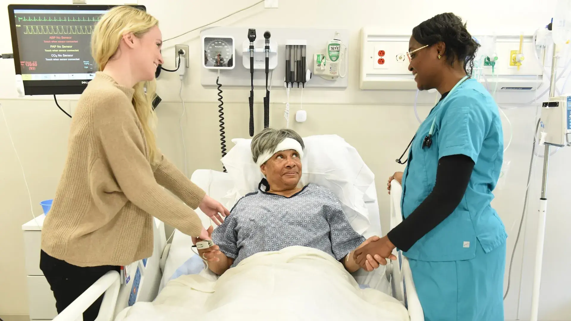two woman hold the hands of an older woman in bed with a hospital gown n