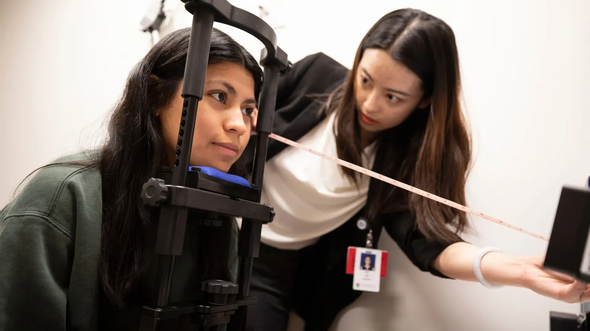 woman rests chin on bar device while another woman measures the distance from her face to a camera