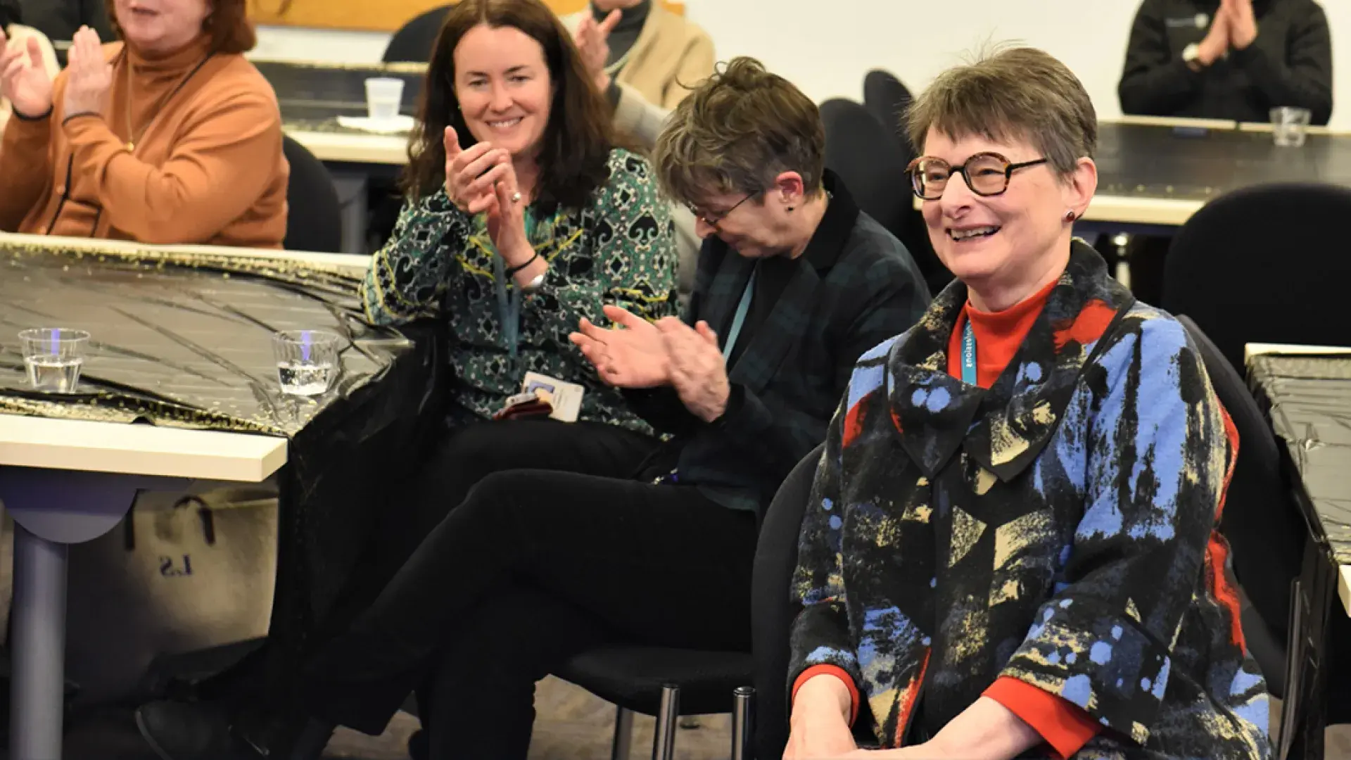 woman with glasses seated at a table smiles broadly as people clap around her