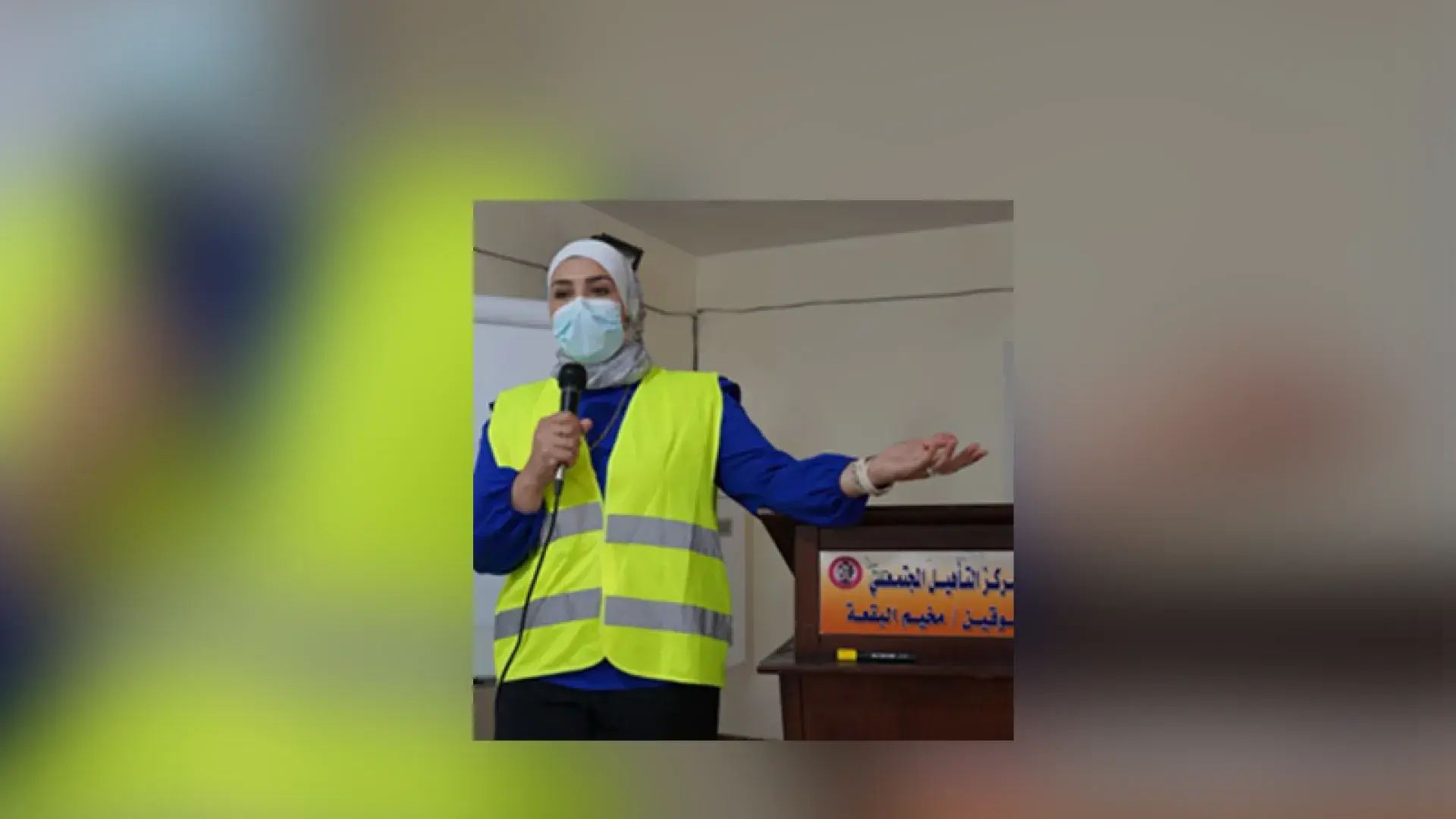 woman in yellow vest and face mask speaks into a microphone