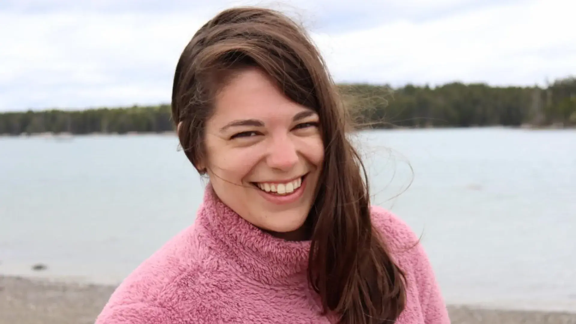 maggie smiles in front of a lake she wears a pink sweater and has shoulder length straight brown hair