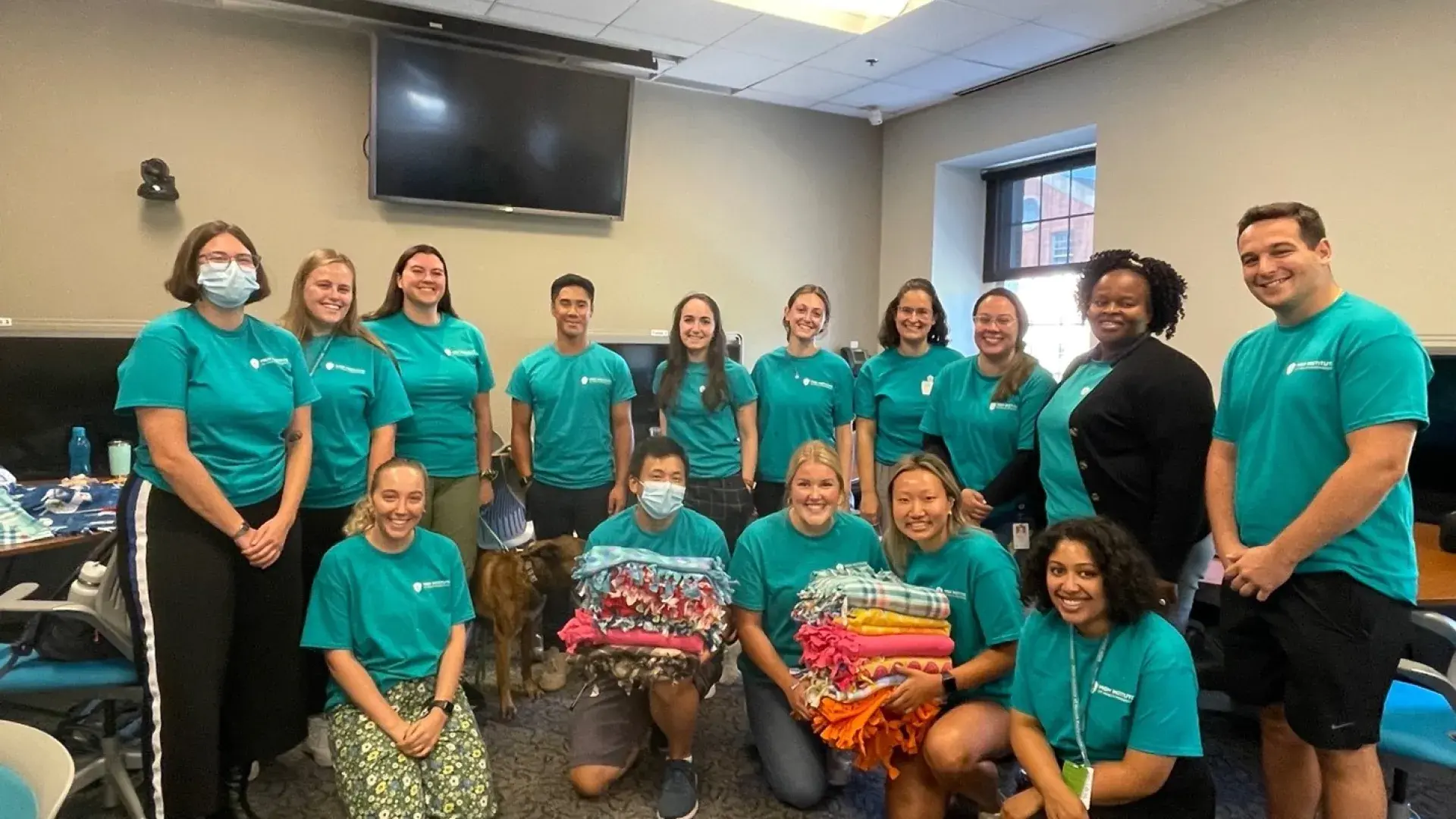 students in matching teal t shirts smile for the camera - two of them hold big stacks of colorful blankets