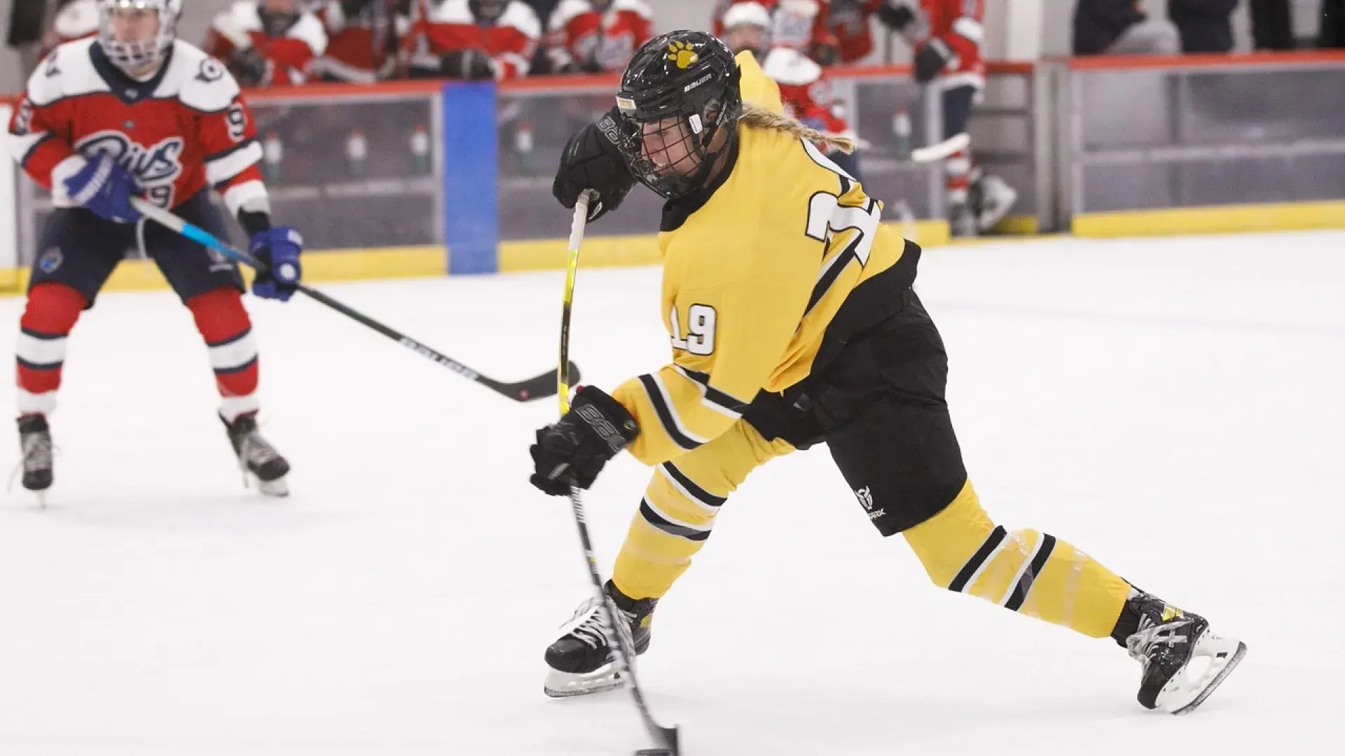 action shot of sammy in her yellow uniform on the ice hitting a puck while a player in red looks on