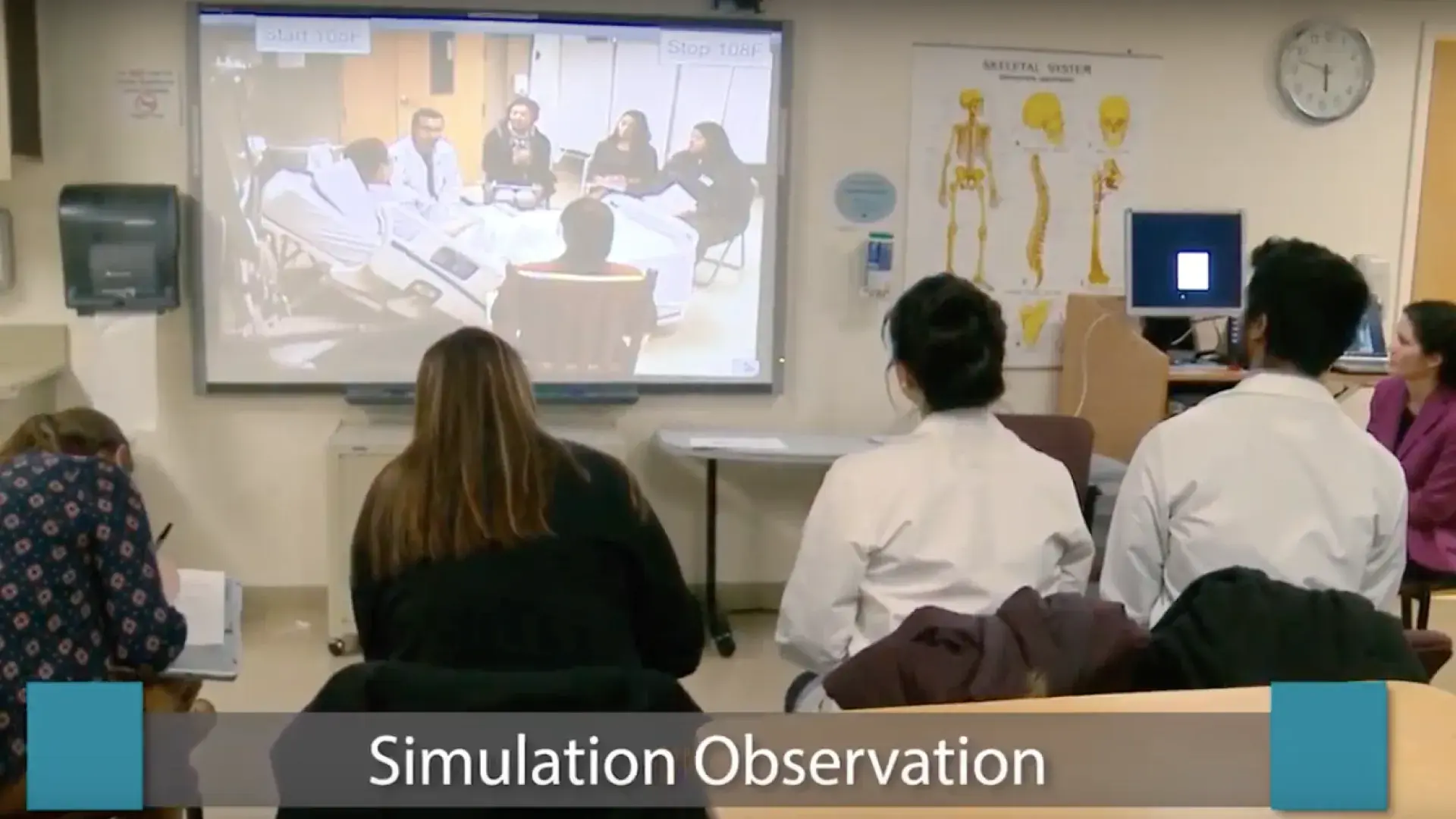 students and faculty sit in front of a screen where they watch other students speak to someone in a hospital bed