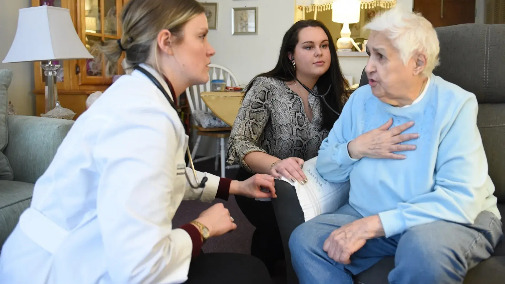 woman in white lab coat squats to be eye level with an elderly woman in an arm chair in her living room