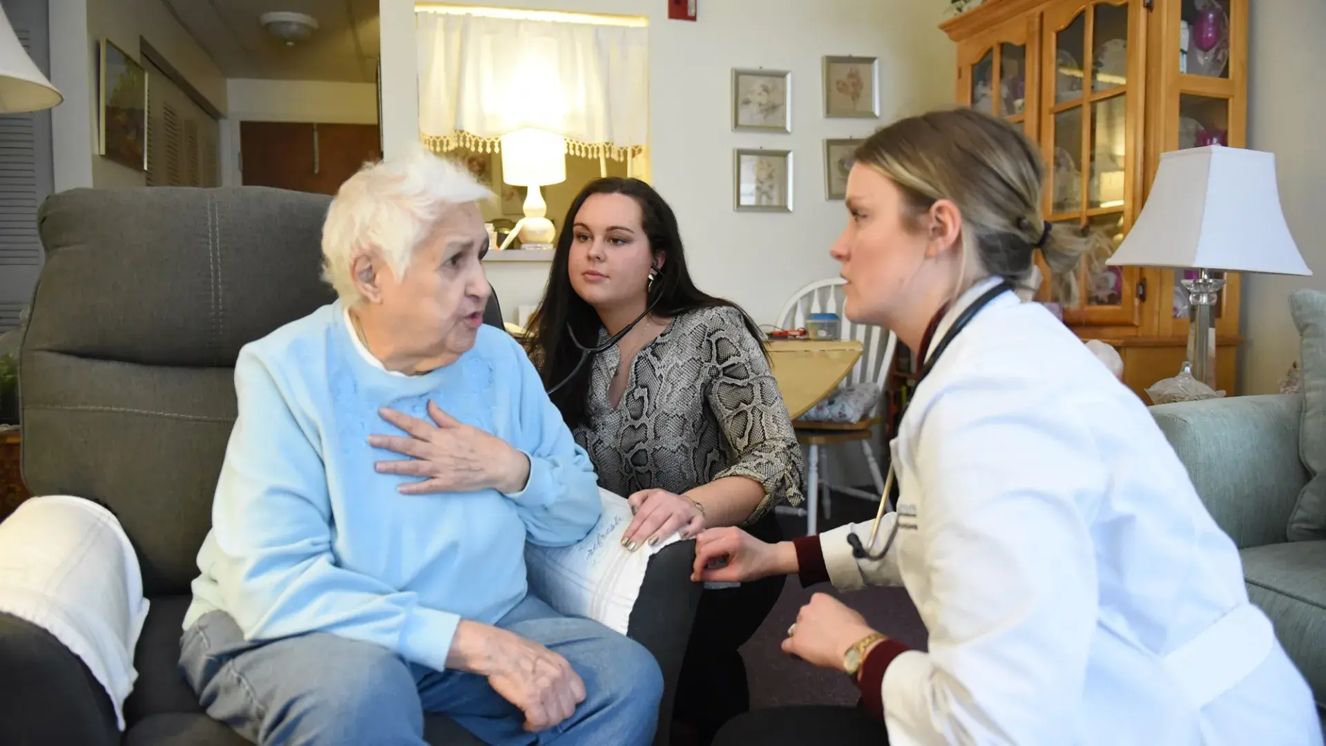 in a living room, an older woman with white hair puts a hand to her chest as two younger females crouch down to speak to her