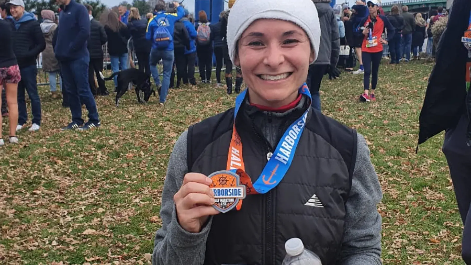Emily stands in front of a big sign that says "start" she wears a winter hat and holds a medal
