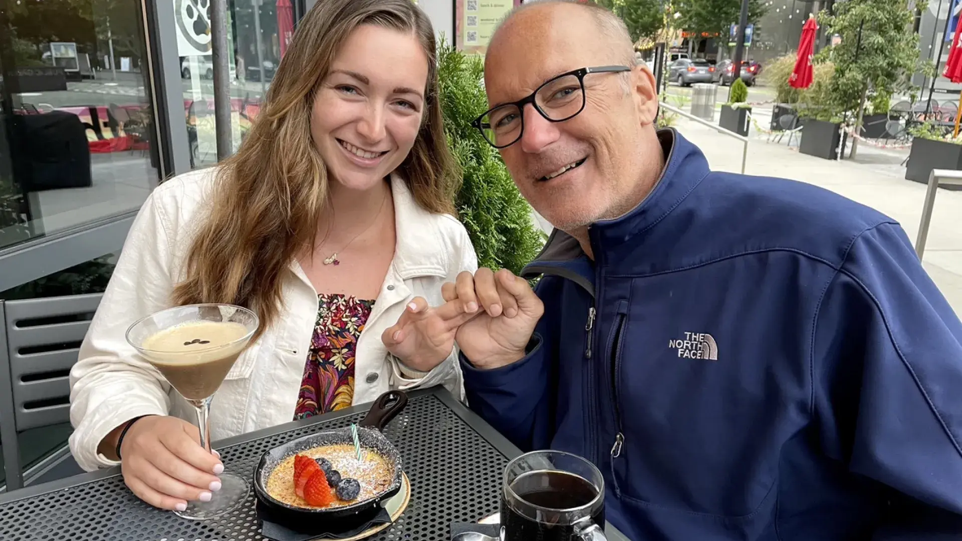 cailin and her dad sit at a cafe table outside with drinks - they smile and hold hands