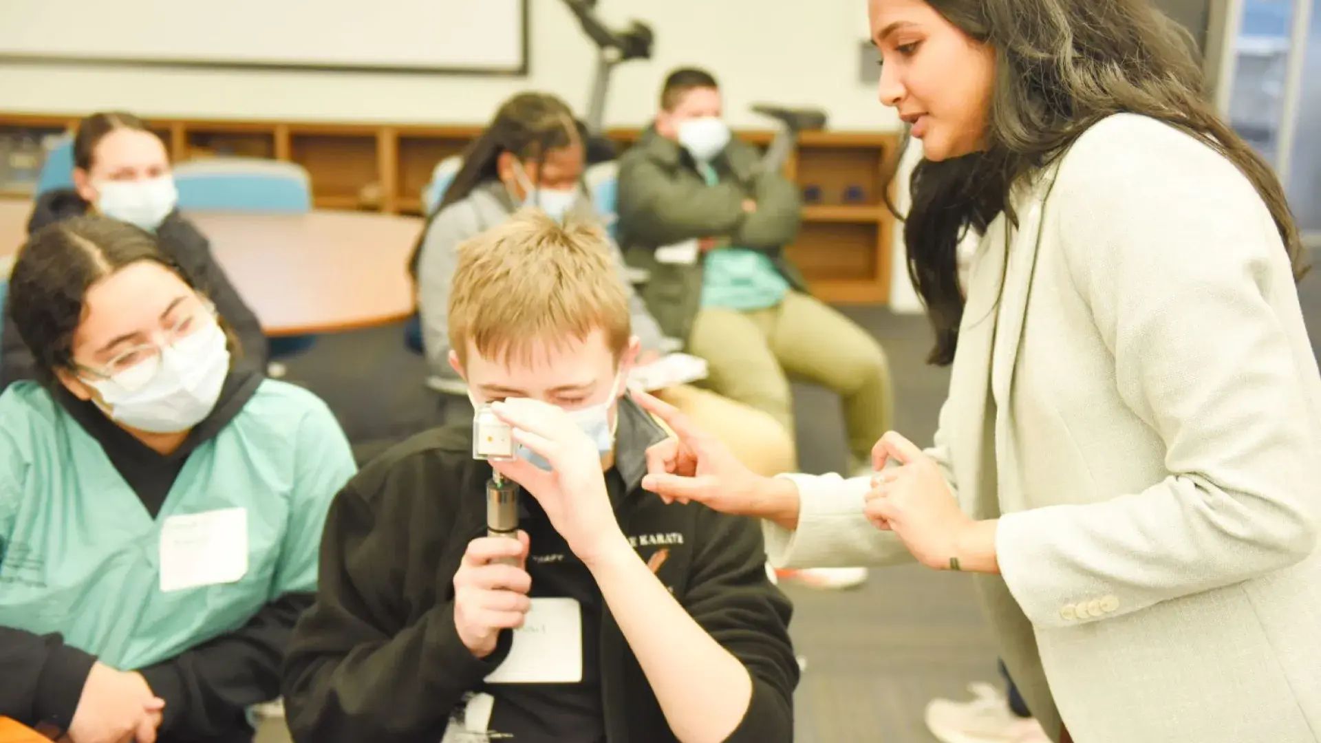 woman in blazer points at a cylindrical device that a man is holding to his eye