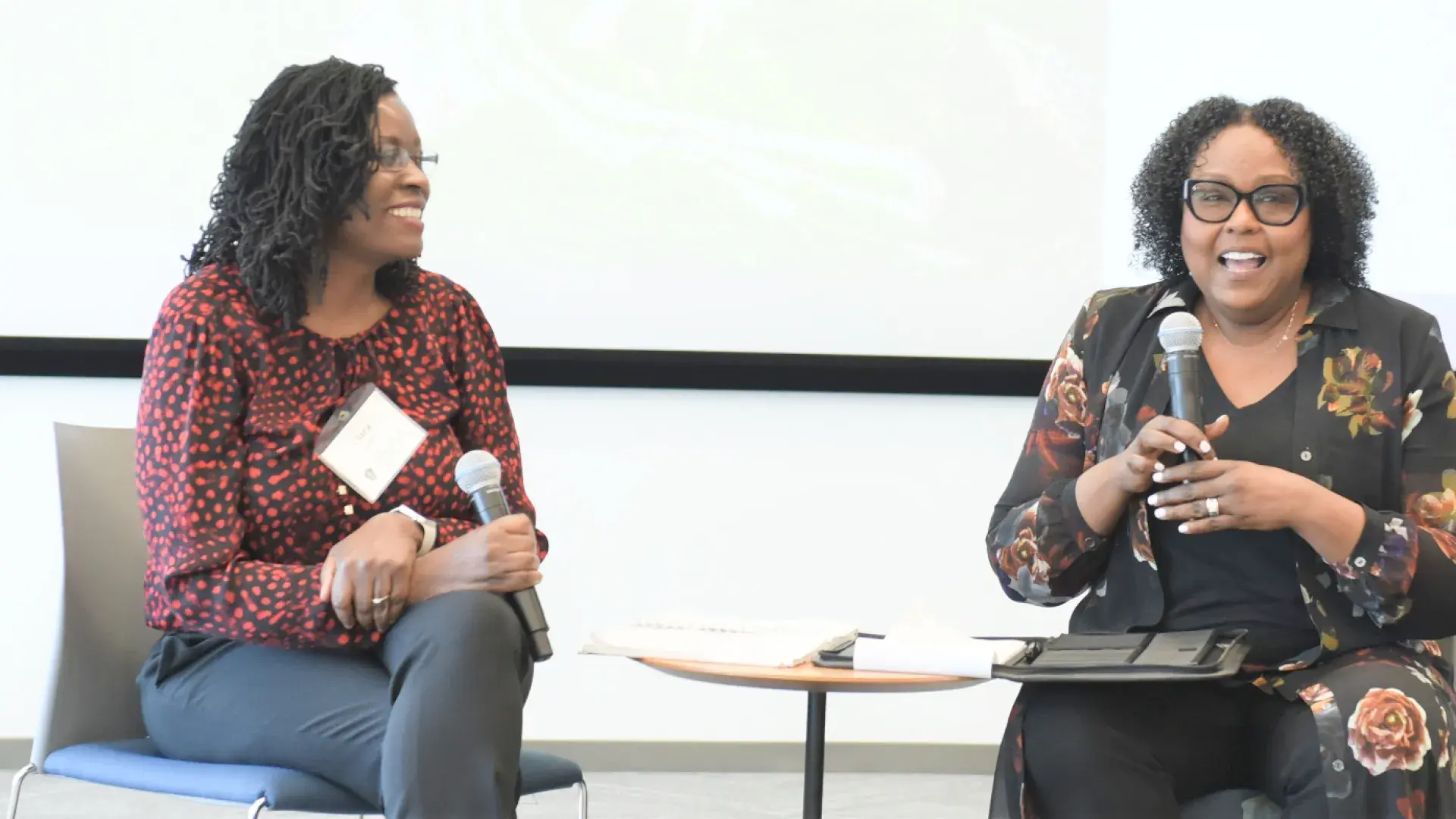 two women of color sit on chairs with microphones