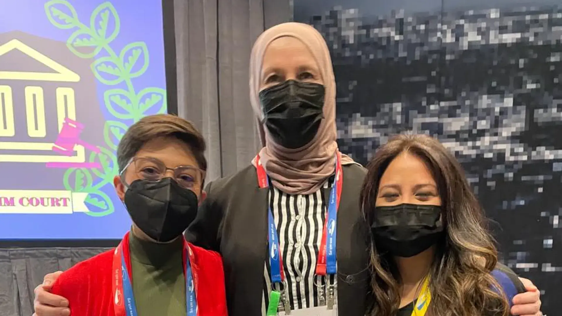 three women stand together in colorful outfits, they wear black face masks