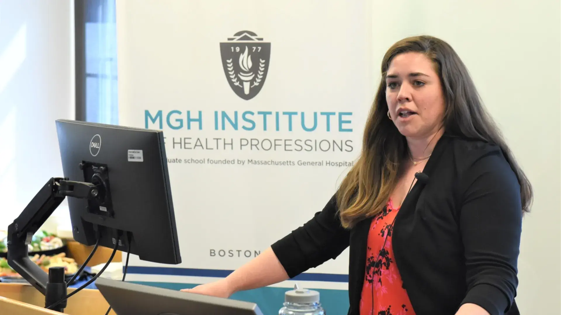 jessica stands at a podium she wears a red top with black flower print