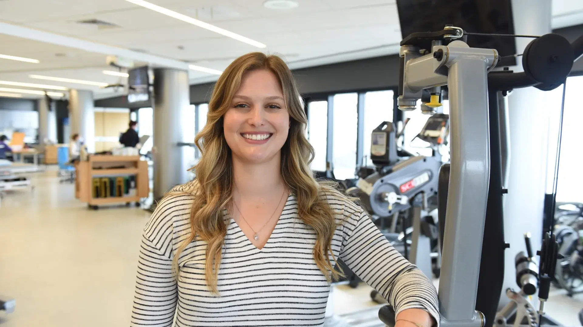 Tarryn has wavy long dark blonde hair and wears a black and white striped top as she leans against a weight machine in a gym