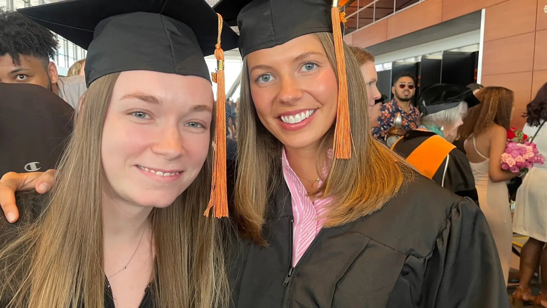 two women wear black graduation caps and gowns, both have shoulder length brown blonde hair