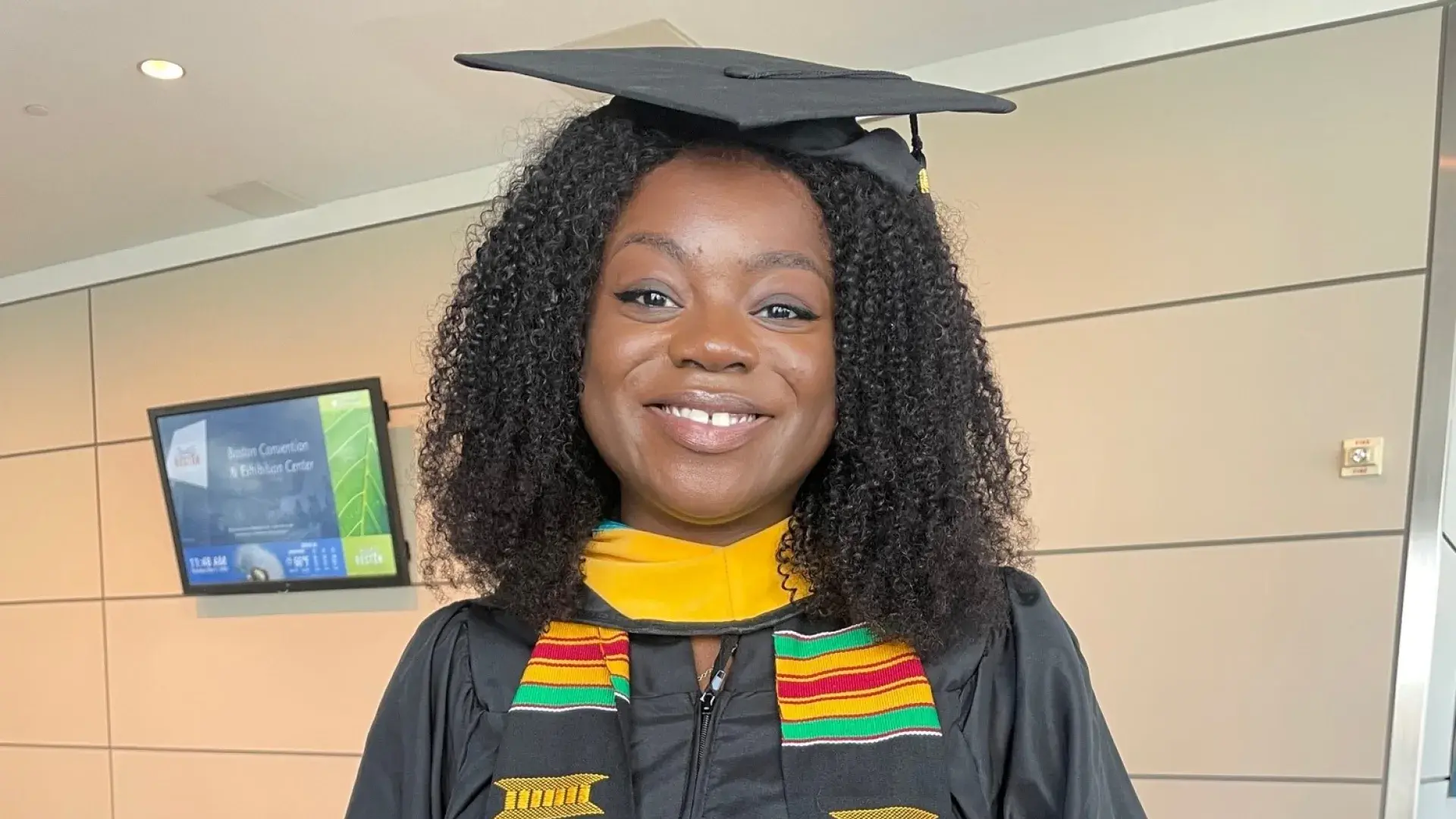 jessica has curly black hair and holds a blue folio. she wears a black graduation cap and gown and green, gold and red striped regalia scarf