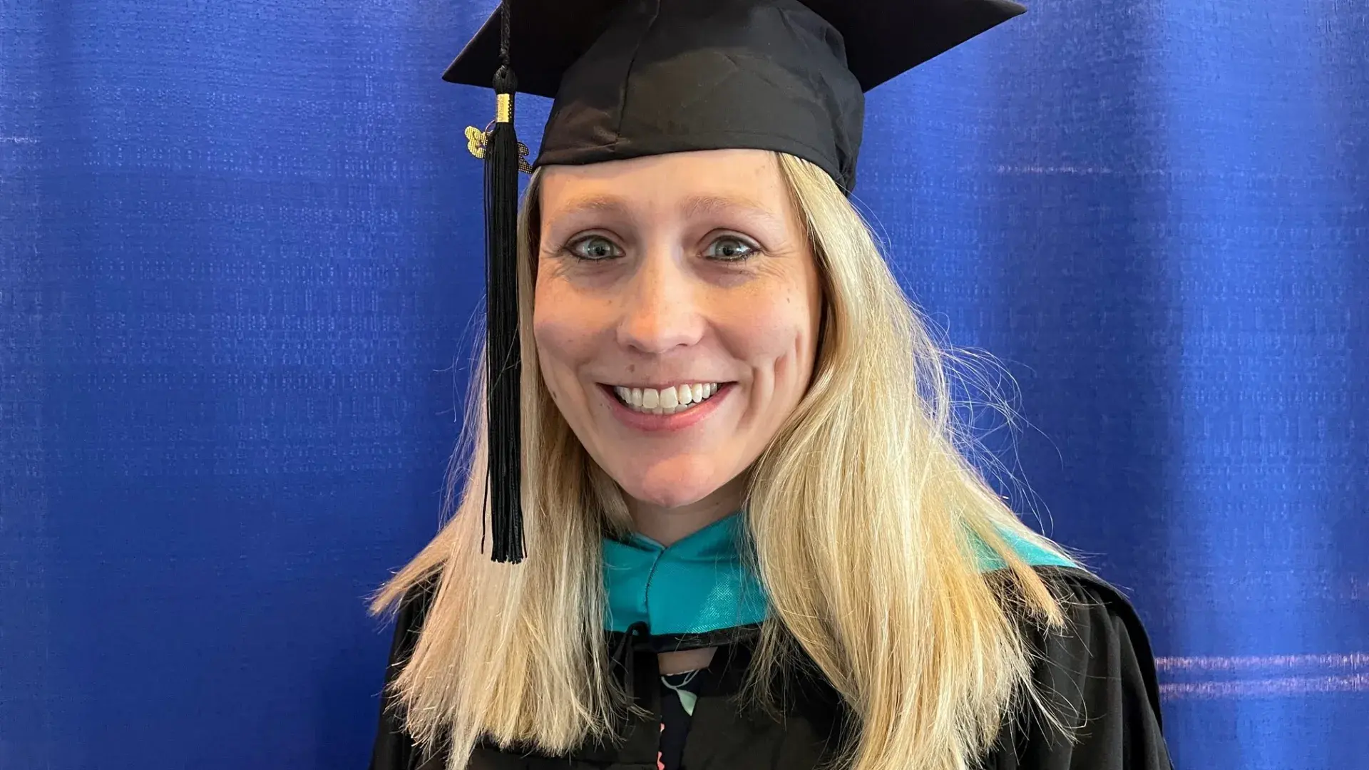 brianna smiles in front of a blue curtain she wears a black graduation cap and gown
