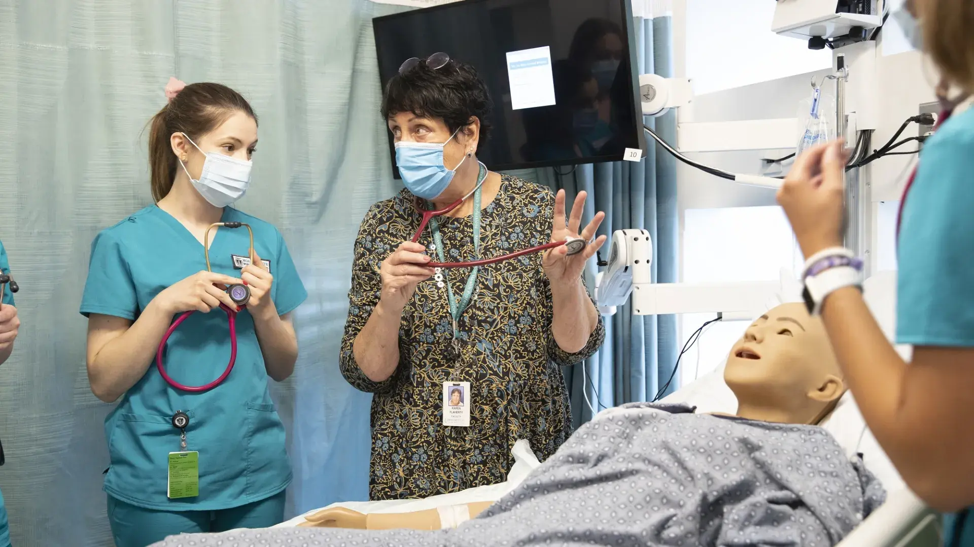 Karen holds a stethoscope and shows a student in scrubs how to apply it to a manikin in a hospital bed