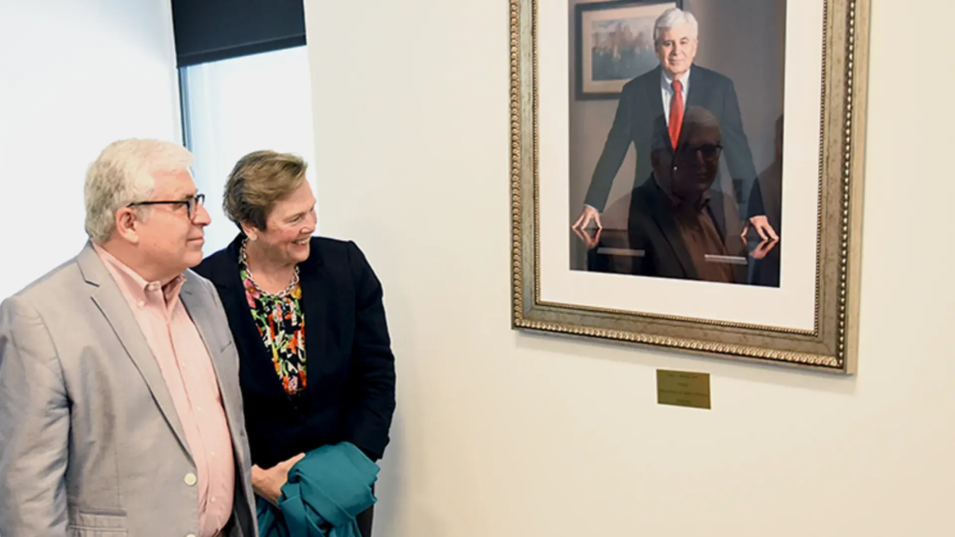 alex in a grey blazer and paula in a black blazer look at a painting of alex in a gold frame on the wall
