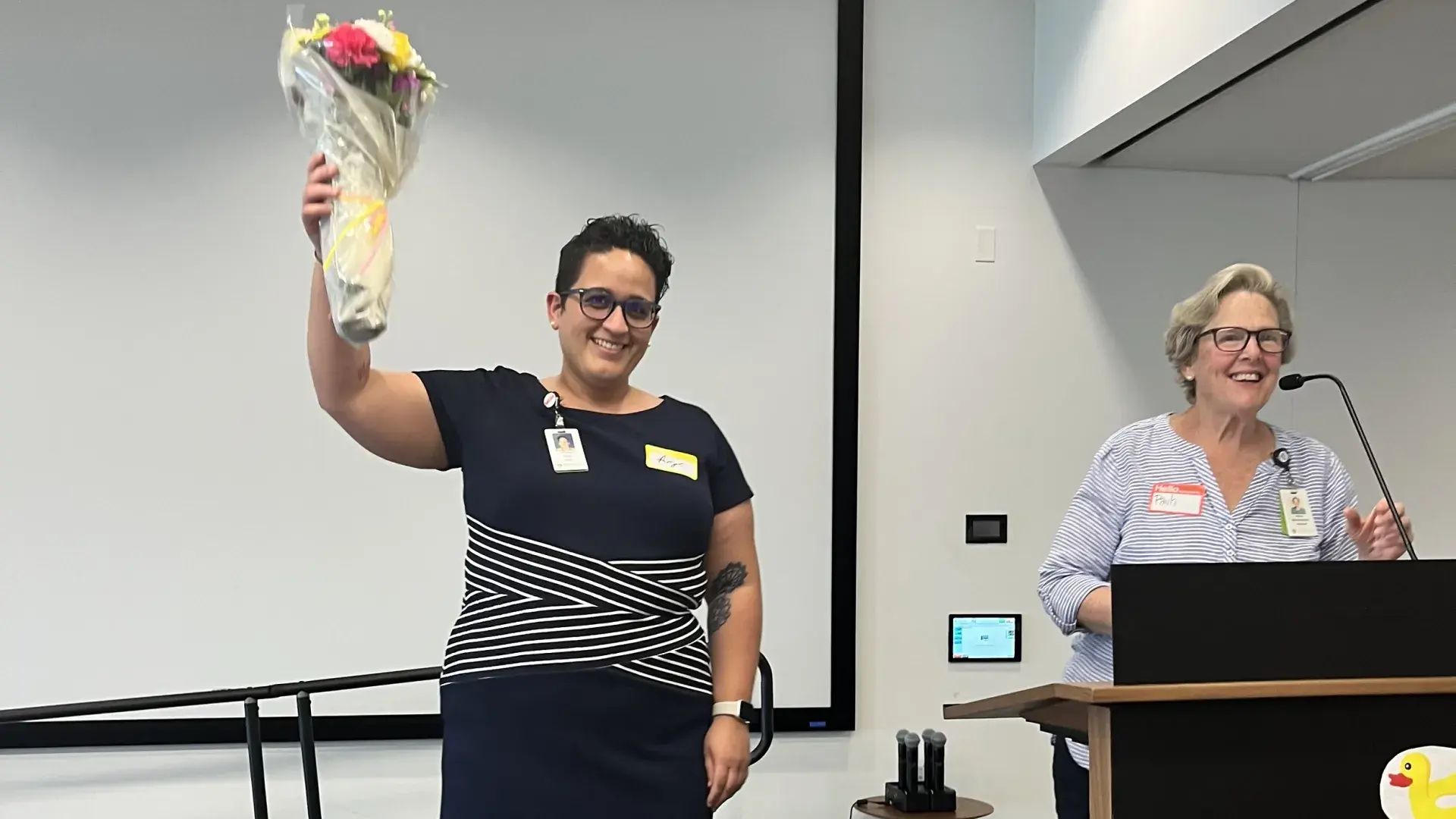 angela wears a black and white striped dress and holds a bouquet of flowers aloft on a stage