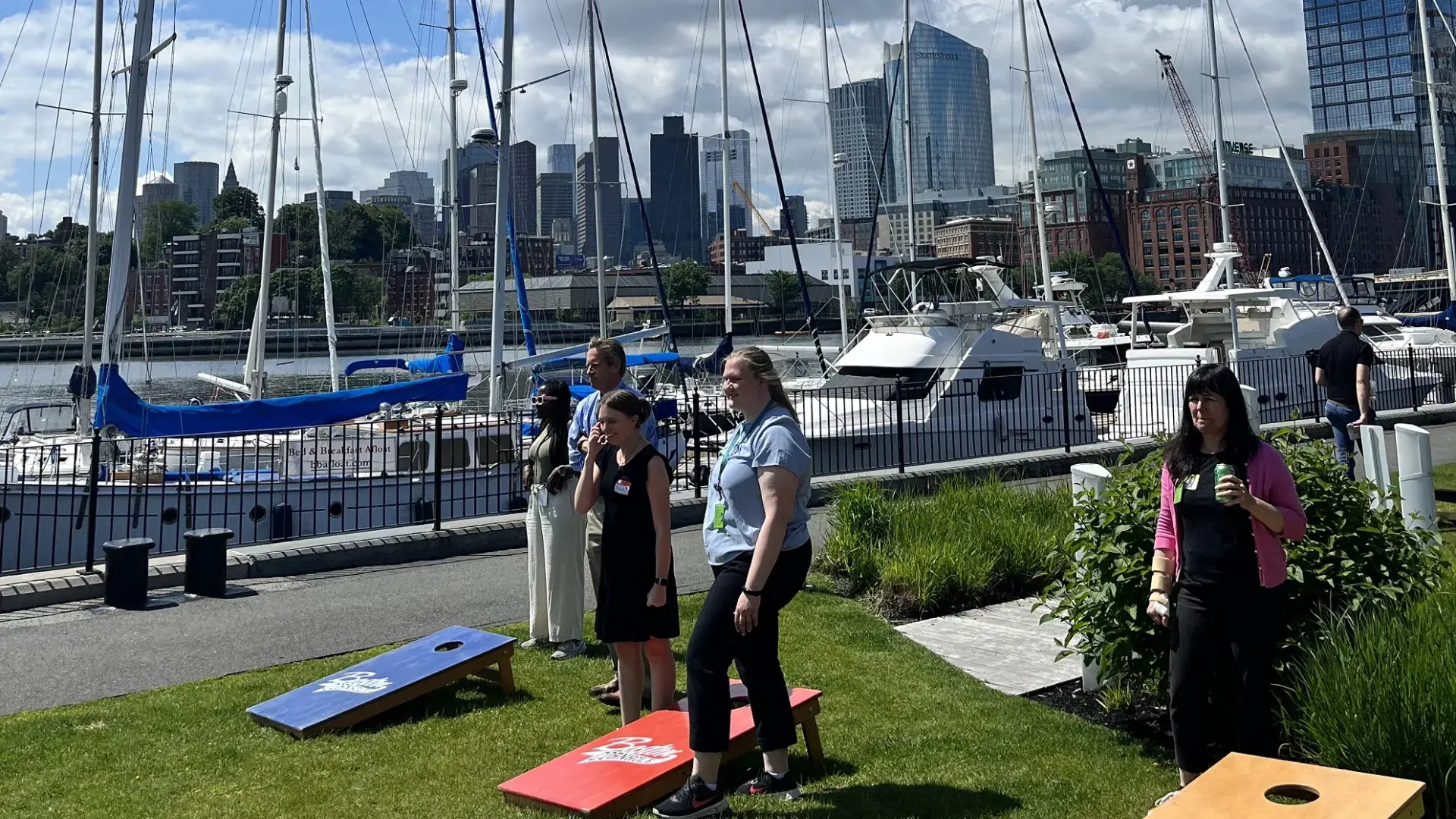 cornhole boards on the lawn in front of a marina with boats and the boston skyline