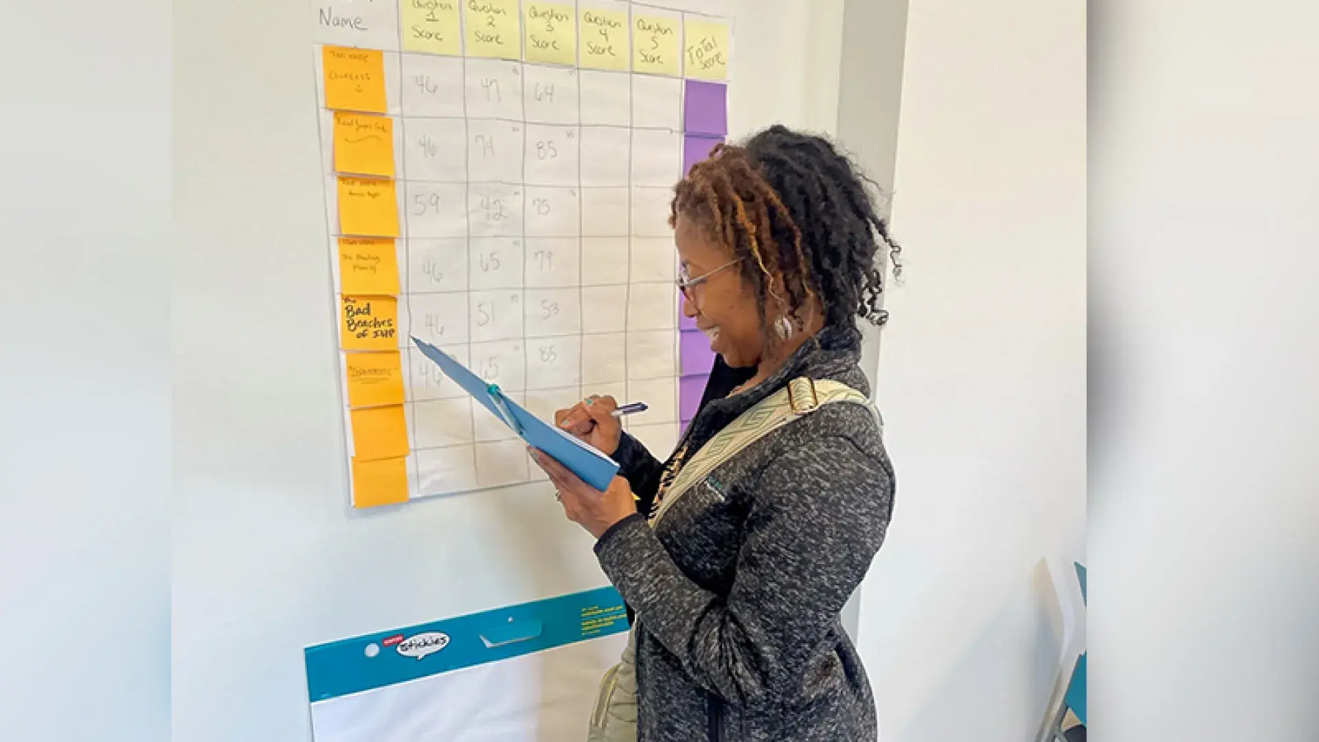 katikie holds a blue folder and stands in front of a board with post it notes