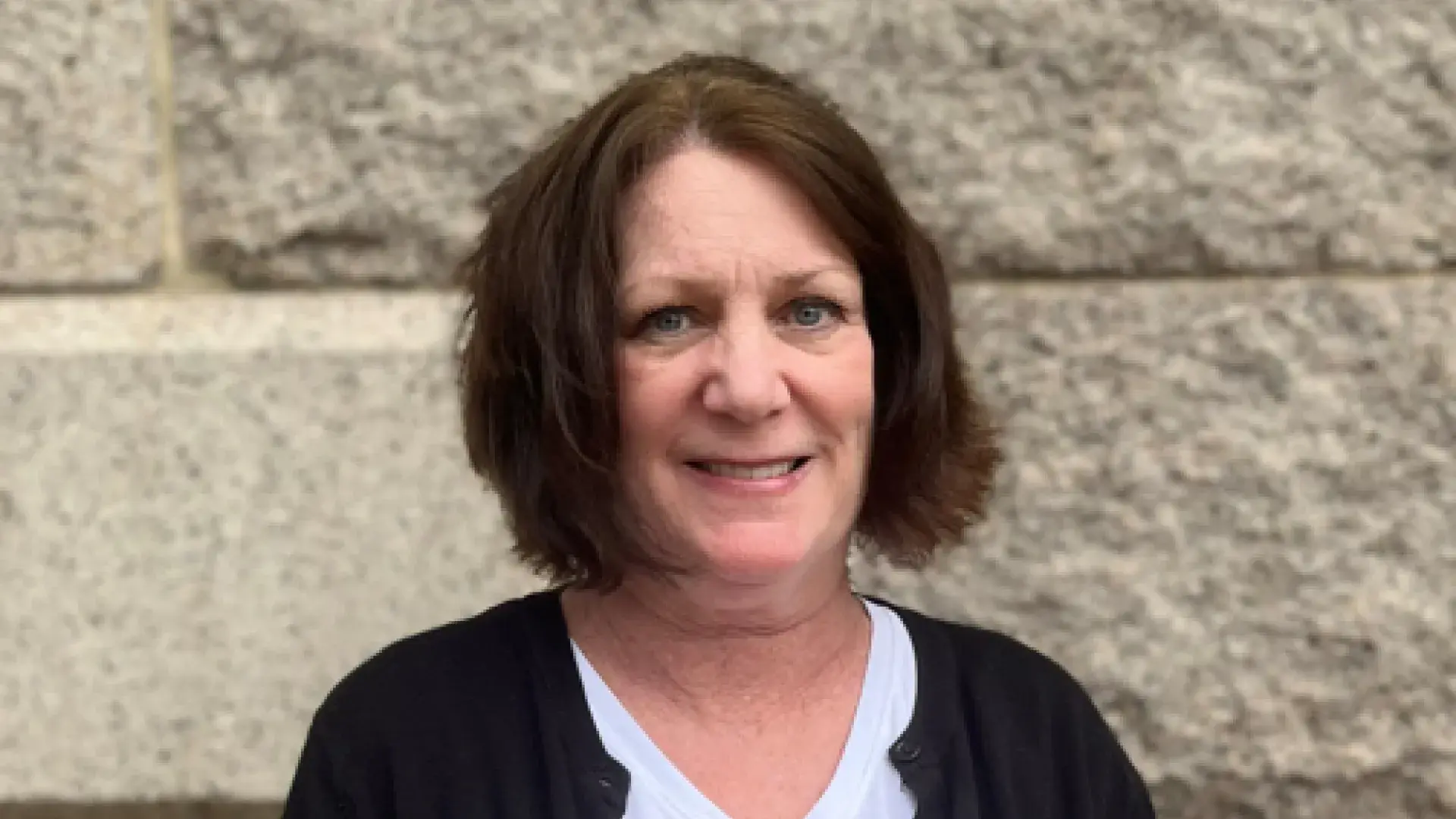 suellen stands in front of a grey brick wall and wears a a black cardigan