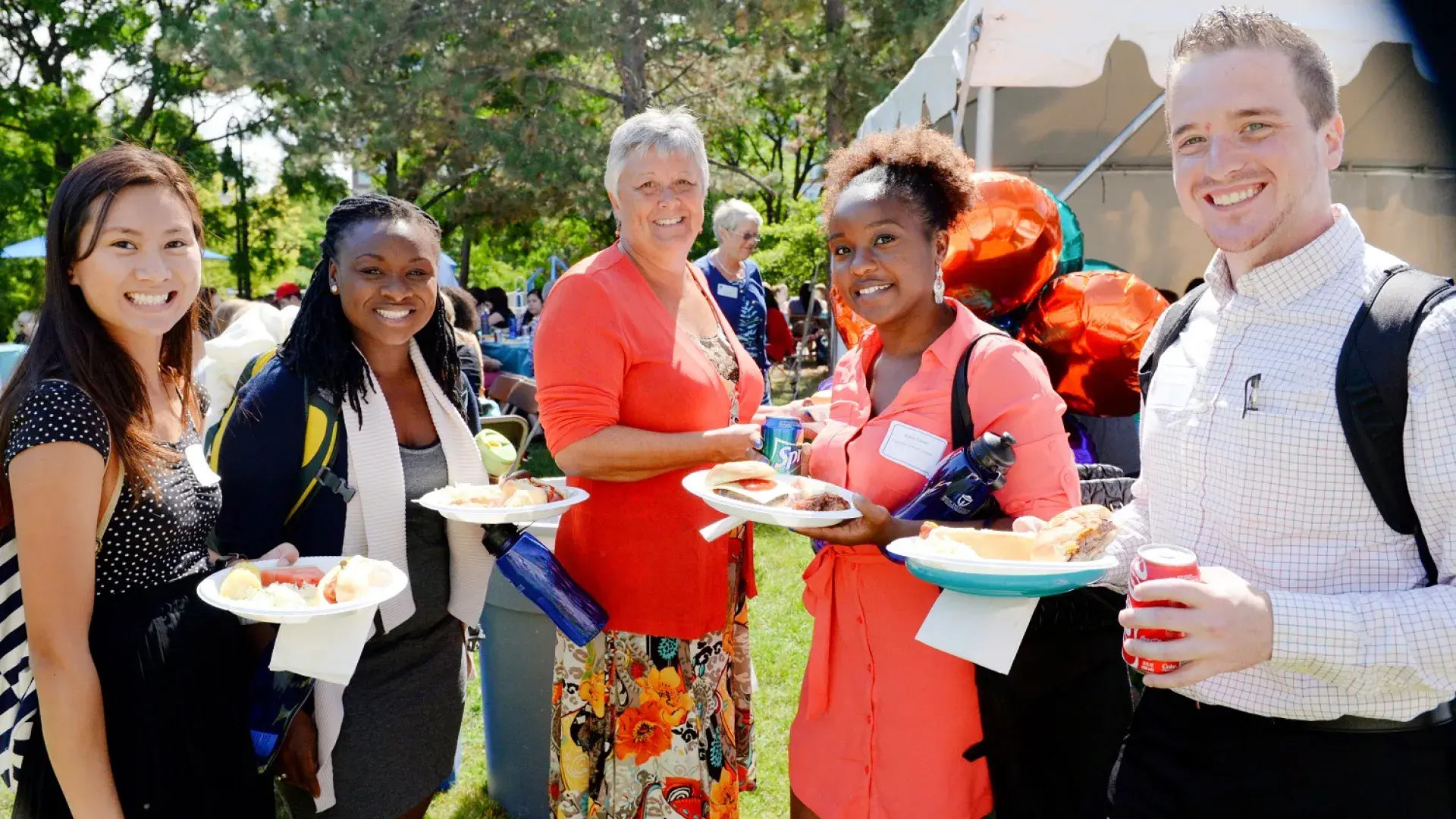 woman in bright orange top with four young-adult looking people in backpacks standing outside on a sunny day with plates of food.