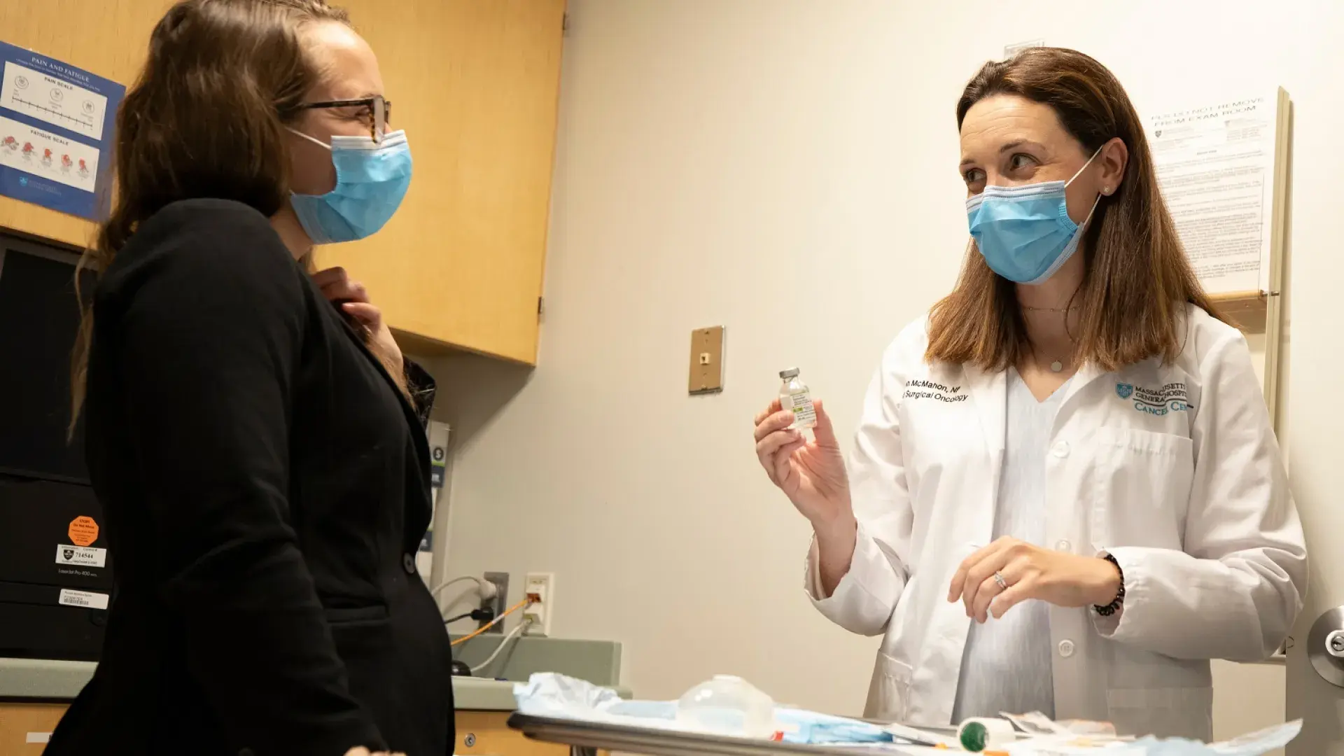person in white lab coat holds up a vial to another person wearing a suit jacket in a doctors office exam room