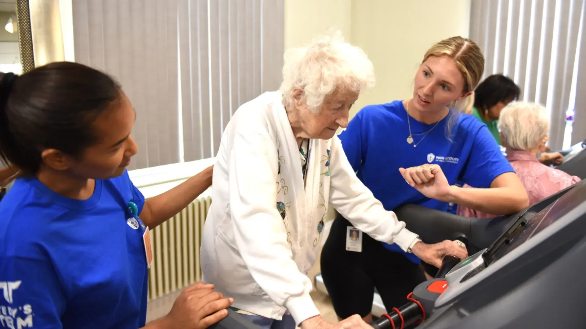 Two young woman work with an older woman walking on a treadmill.