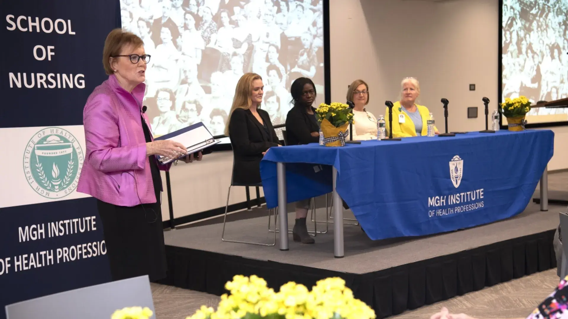Woman standing speaks with 4 people behind her sitting at table