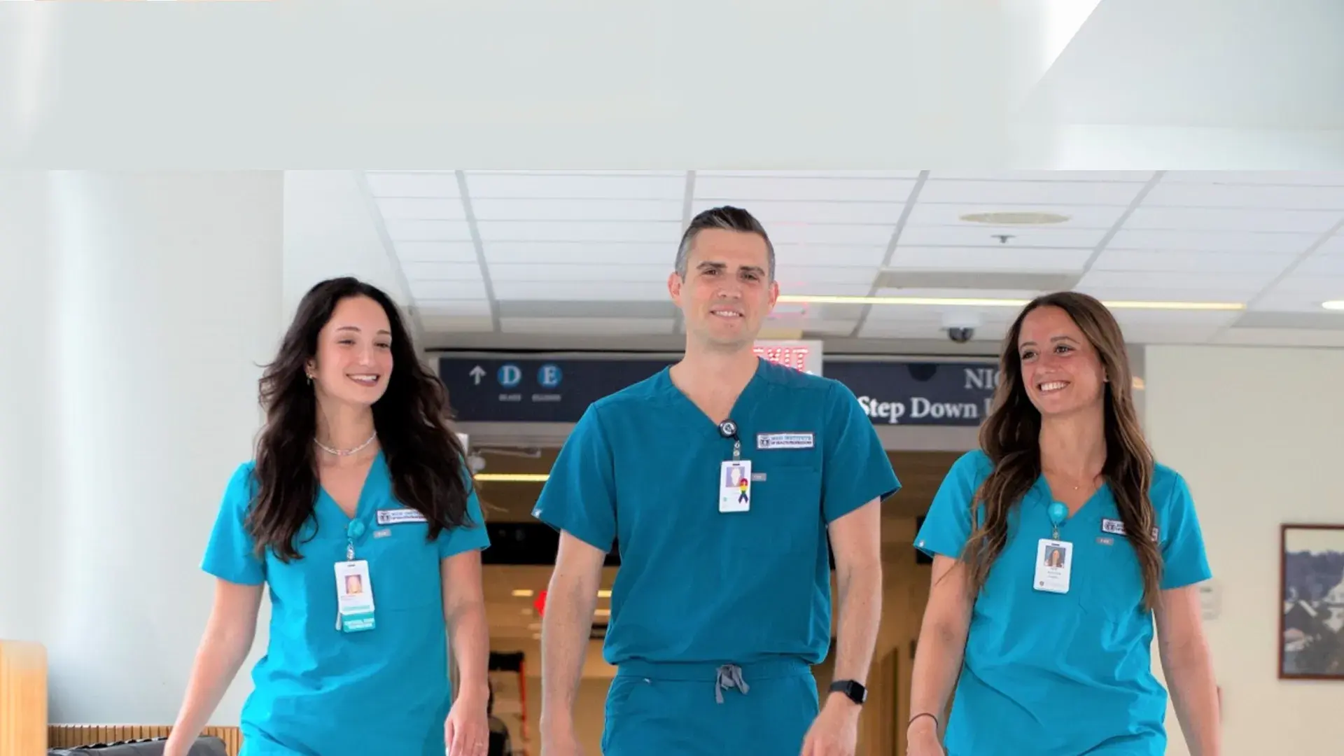 a man and two women in blue scrubs and name badges walk down a hospital hallway (step down unit sign in the background)