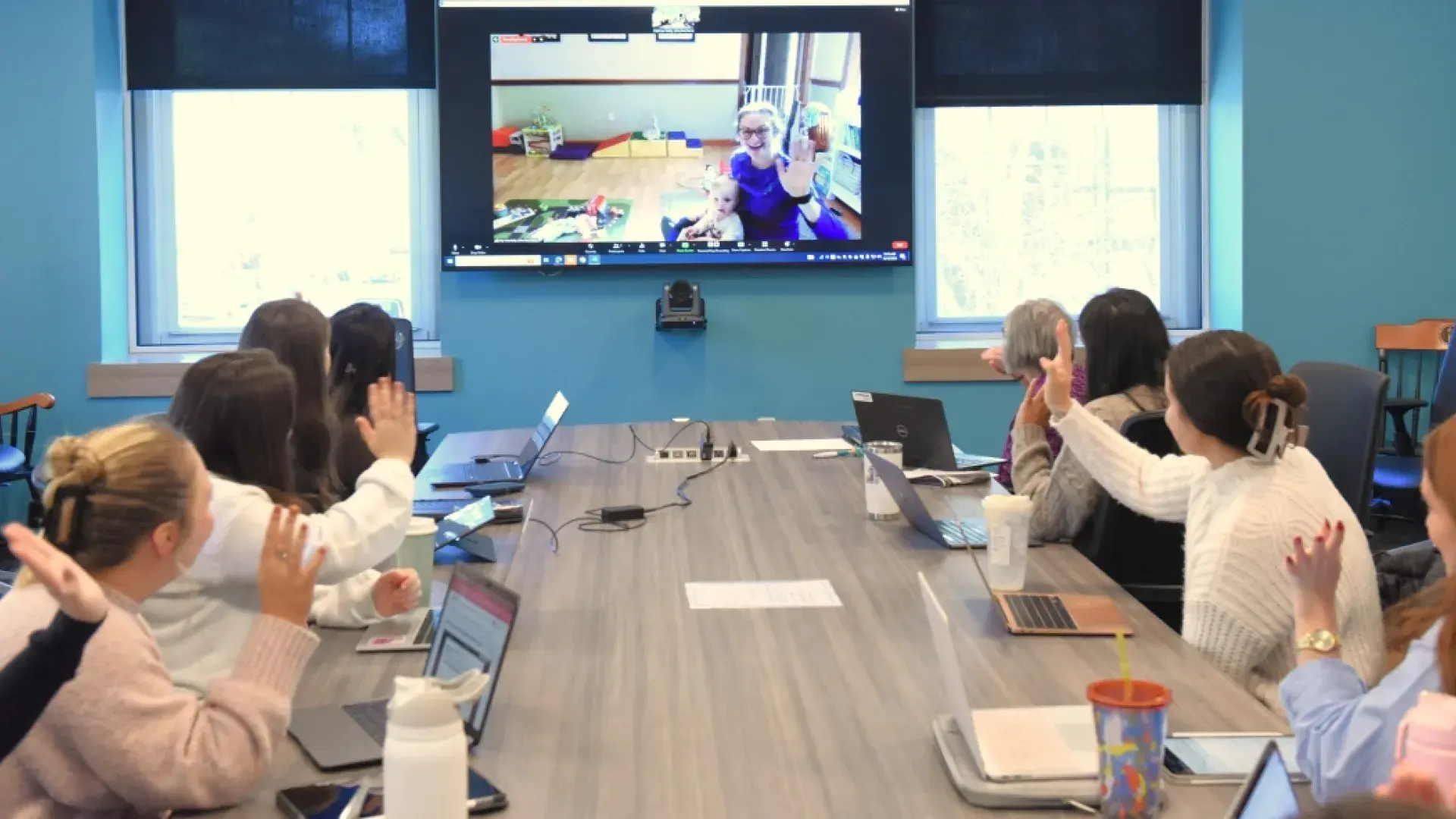 People around a table waving at a wall monitor