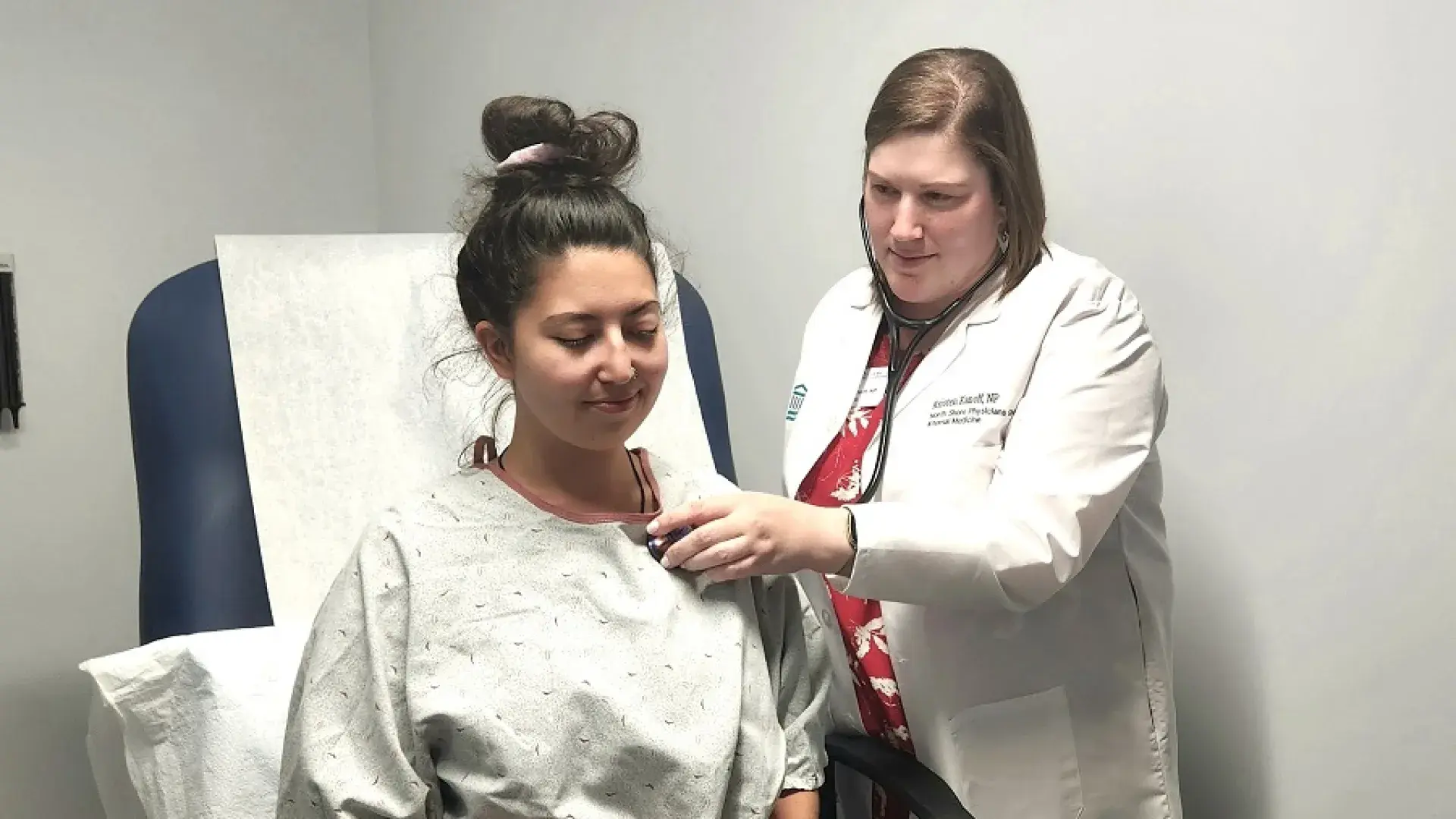 Woman in a lab coat holding a stethoscope to a woman in a hospital gown's back