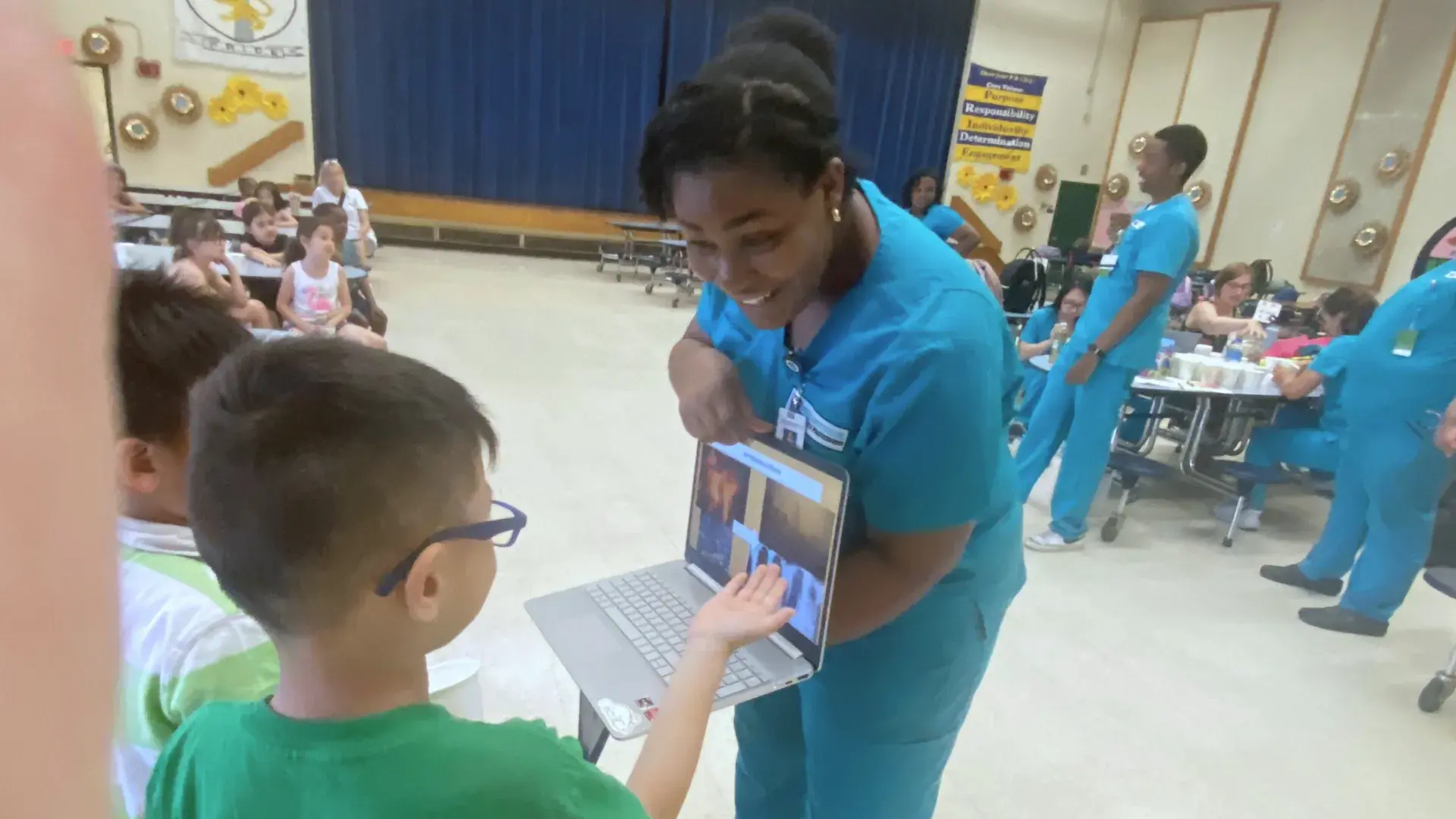 Nursing student in teal shows a pupil something on a laptop
