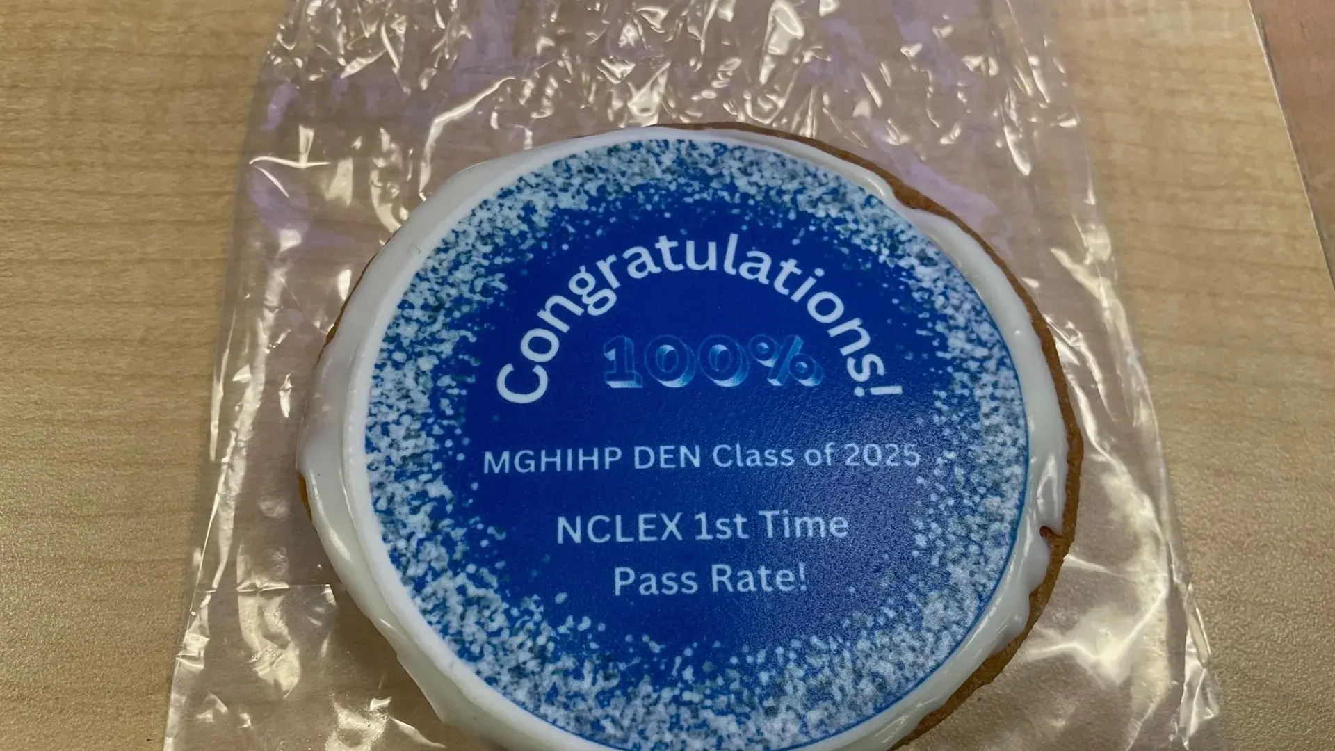 Decorative cookie celebrating MGH IHP DEN Class of 2025's 100% NCLEX first-time pass rate, featuring congratulatory text and blue frosting design.