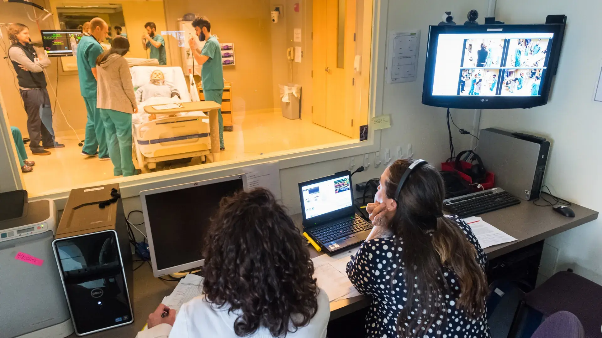 two people sit behind a glass window and watch 5 people in scrubs around hospital bed where a manikin is laying
