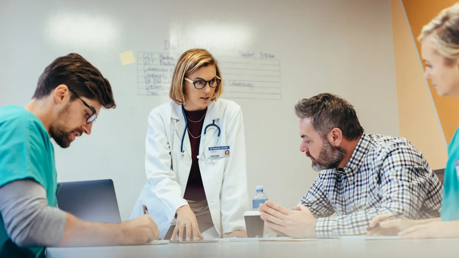 woman in white coat and stethoscope gestures to people at a table wearing scrubs