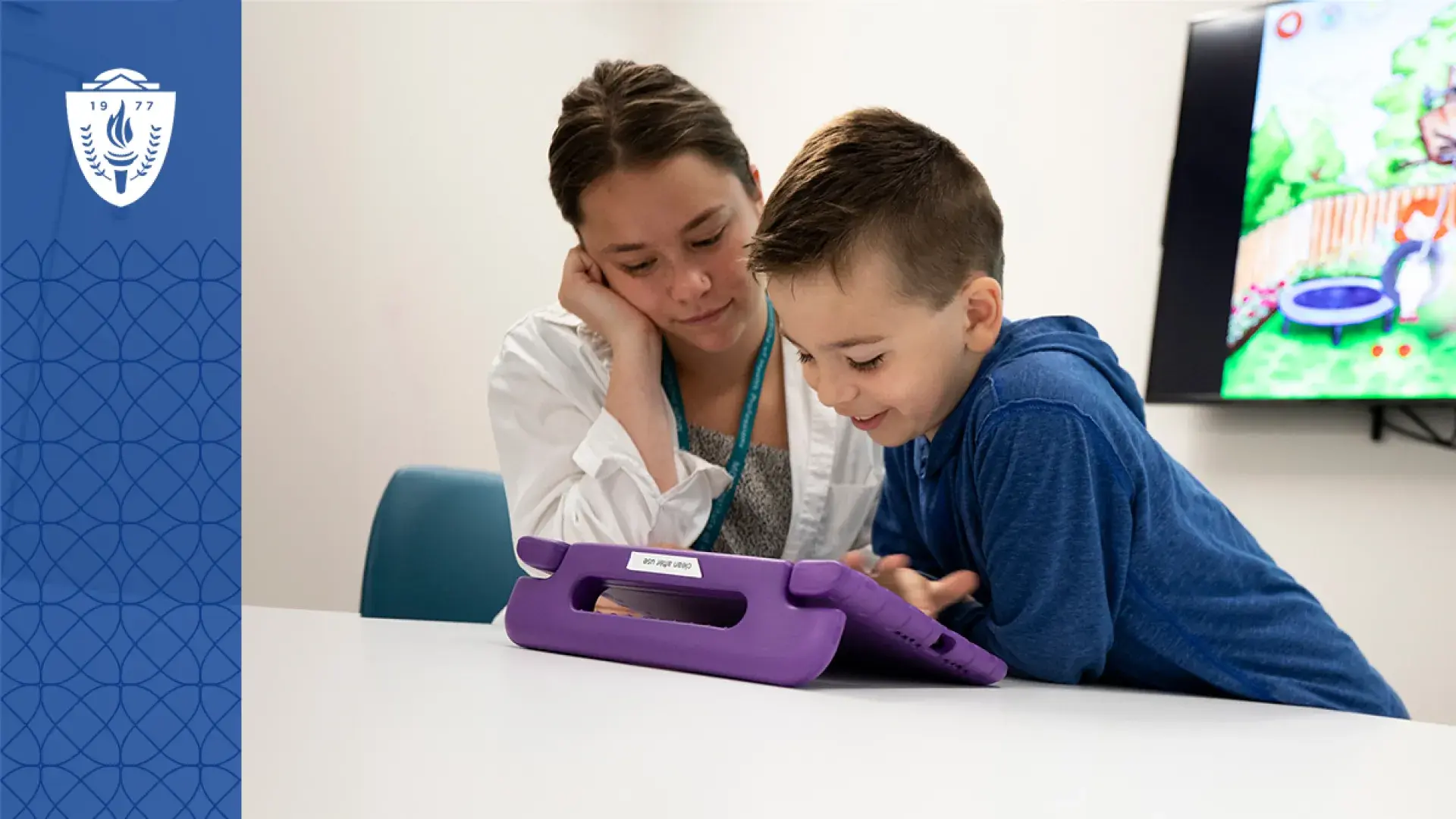 student in a lab coat looks at something on a tablet with a little boy while a colorful cartoon is on the TV in the background