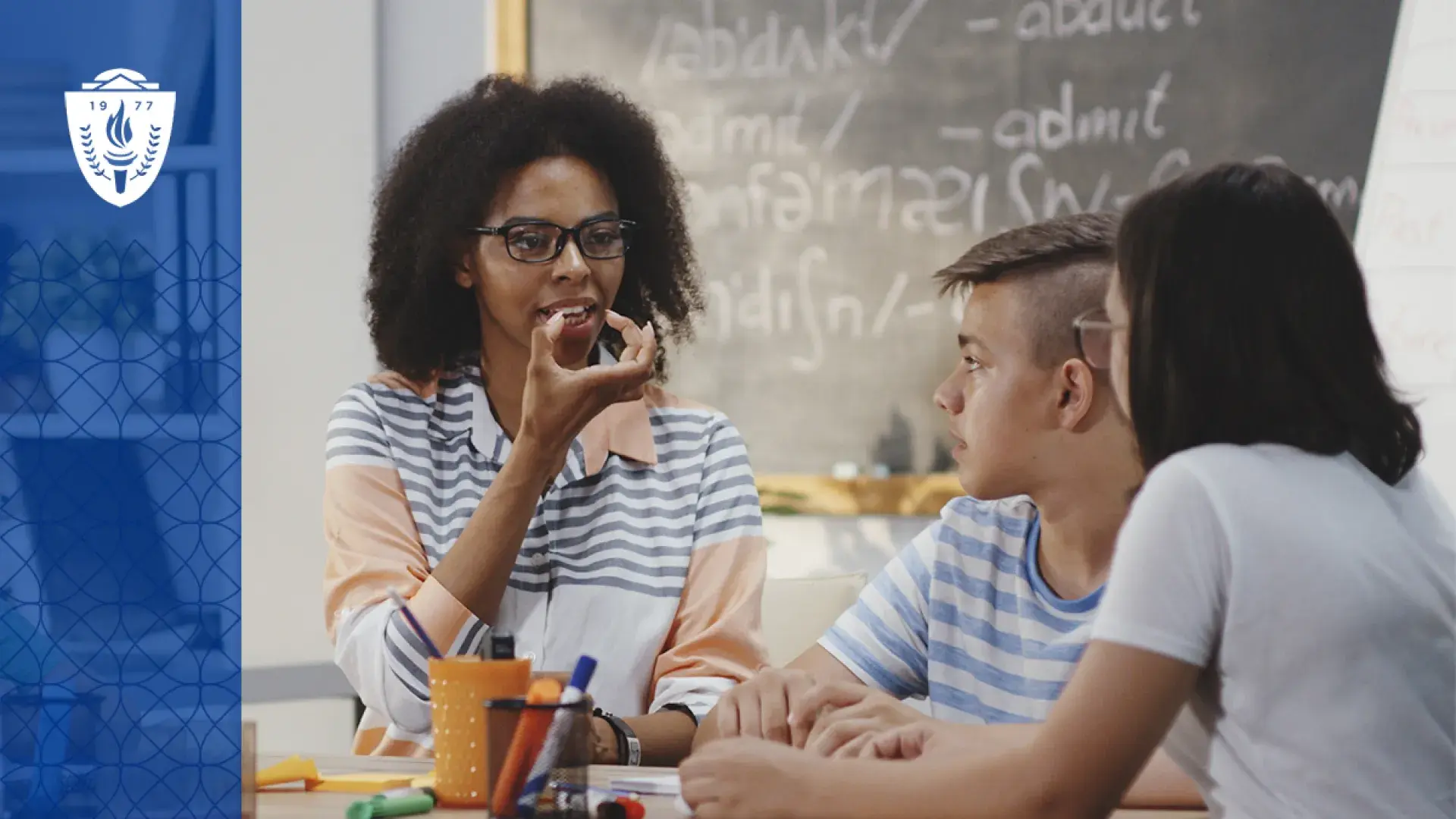 a woman in a classroom holds her hand to her mouth to show how to form a word to two younger students
