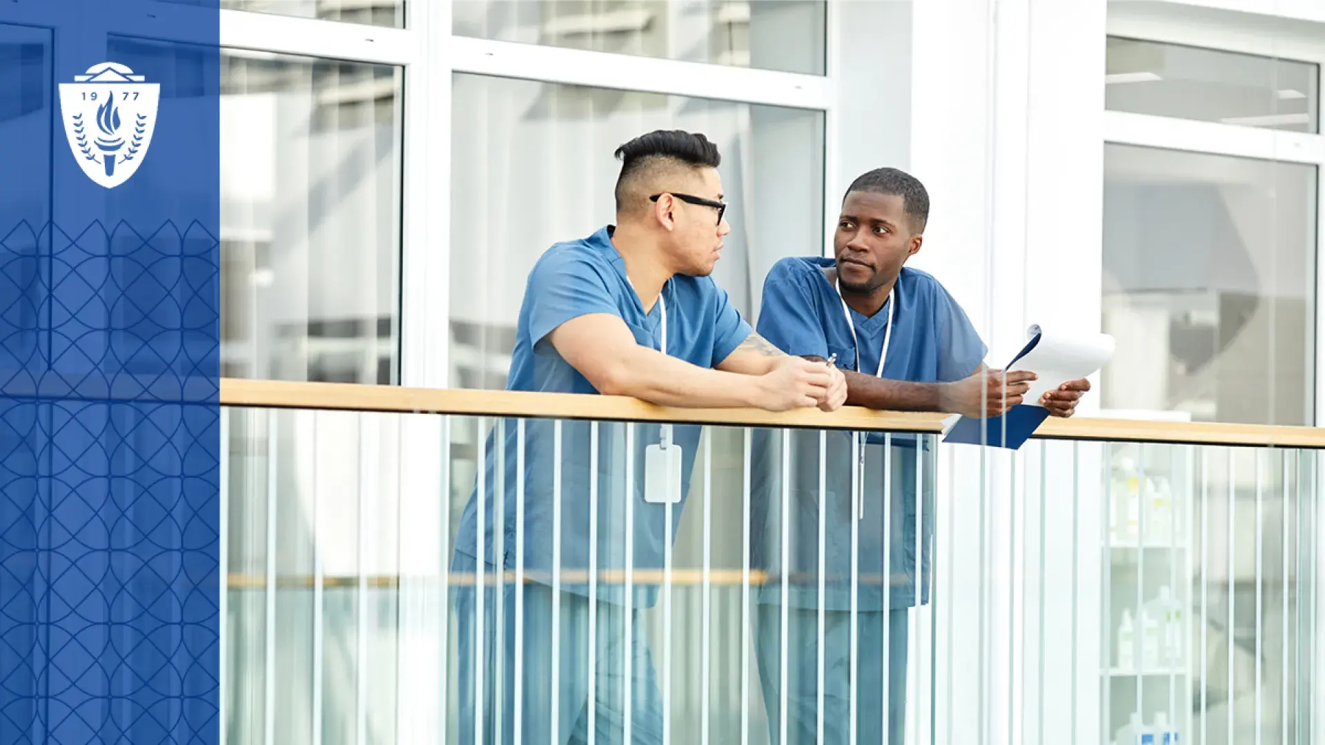 two men in scrubs lean on a hospital railing and converse with each other