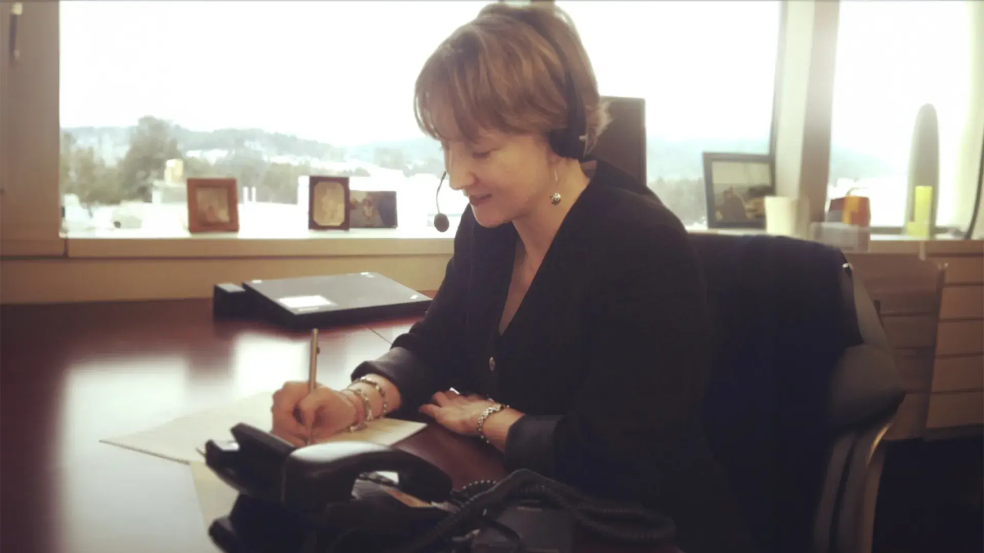 kathy sits at a desk with a headset on and takes notes on a sheet of paper