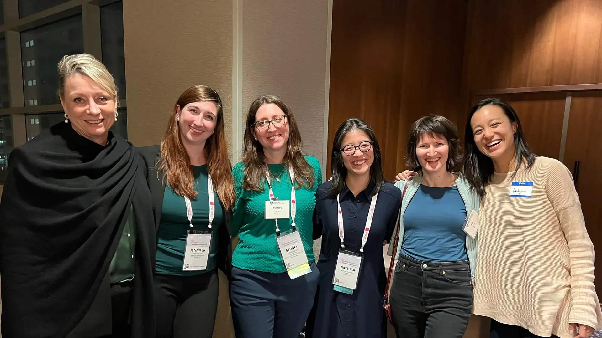 six women stand with their arms around each other, they are wearing rectangular badges on lanyards around their necks