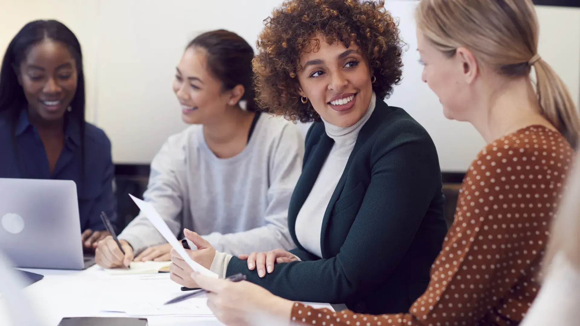 four women in business casual attire sit at a table and discuss paperwork
