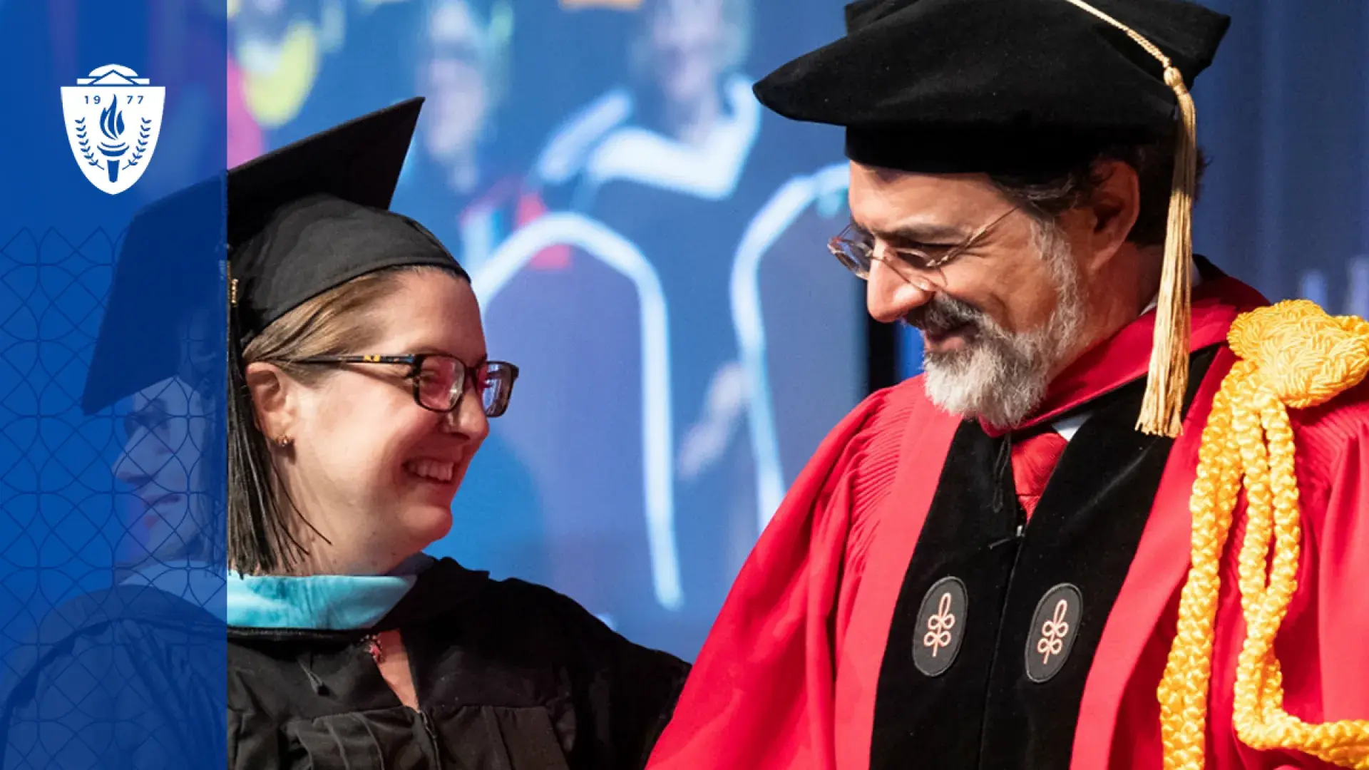 professor in graduaiton regalia (cap and red gown) smiles at a woman graduating who is in blue regalia