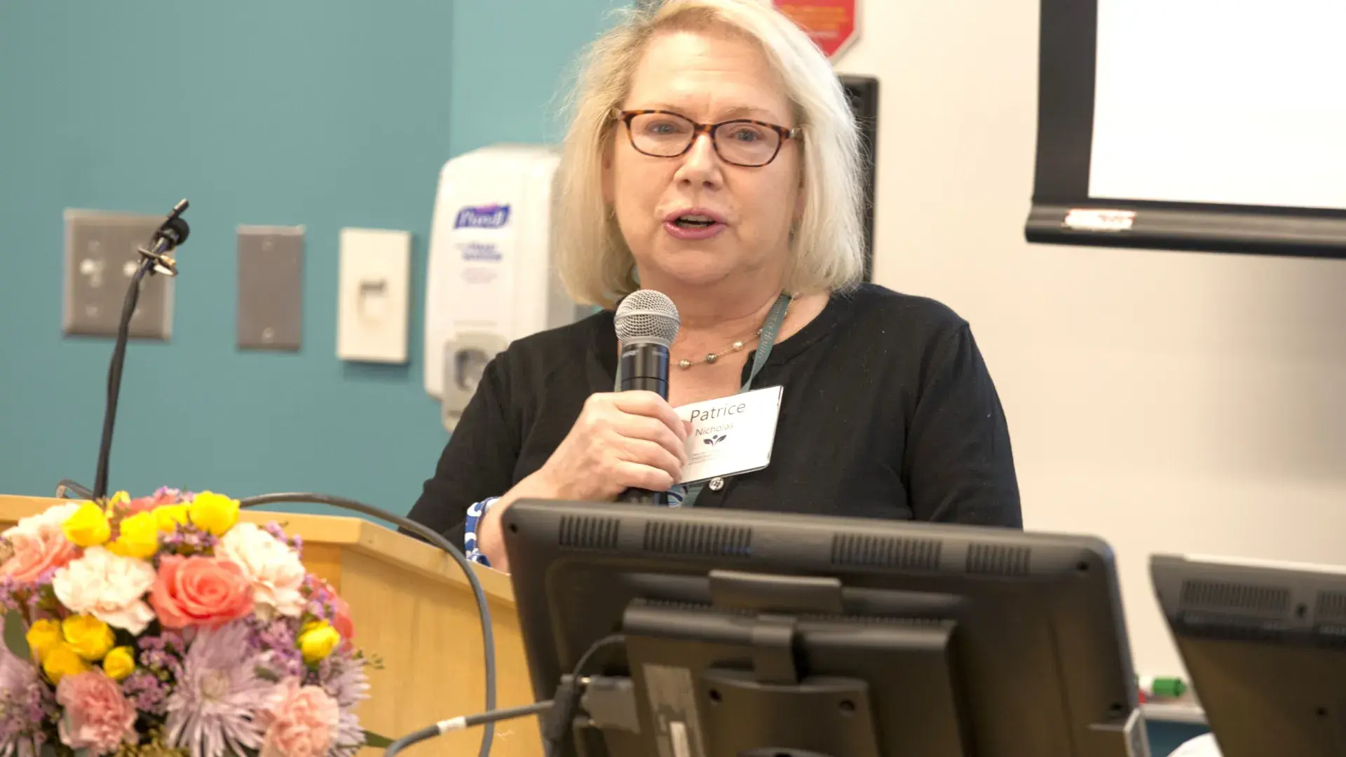 A woman holding a microphone standing next to a computer screen and flowers