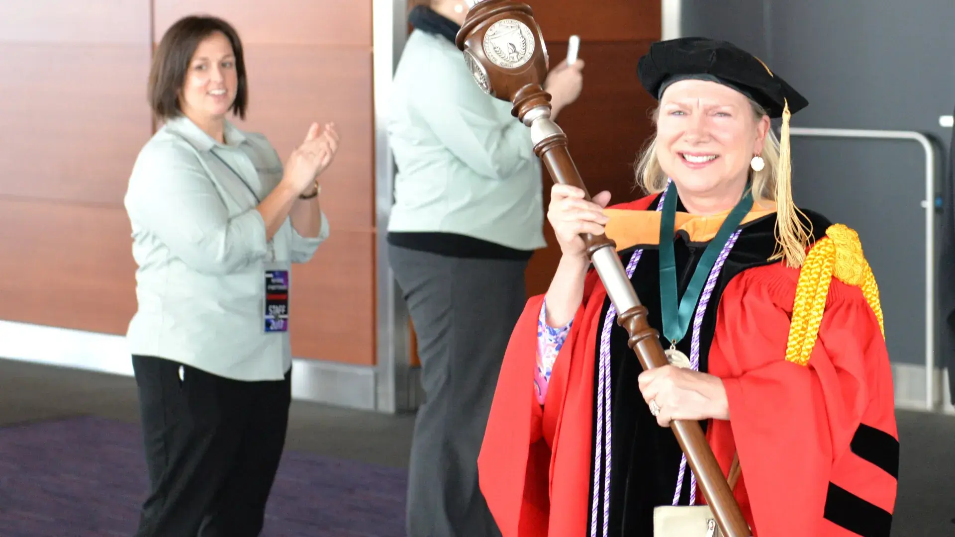 A woman in commencement attire holding a large scepter 