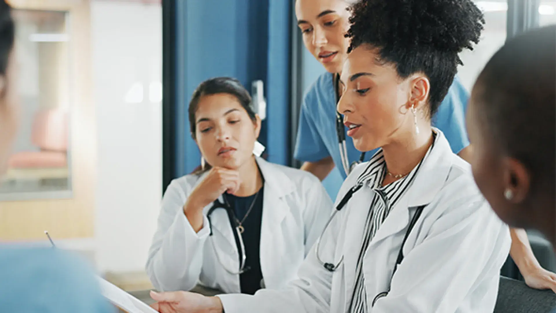 woman in lab coat and stethoscope showing others around the table something on her ipad 