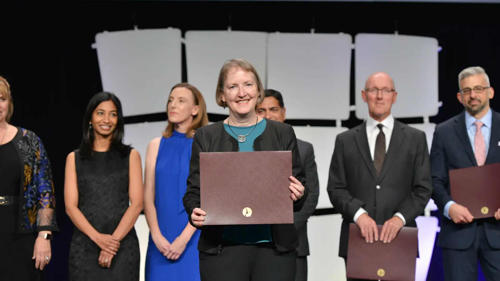 A woman standing on a stage holding an award with a line of people behind her